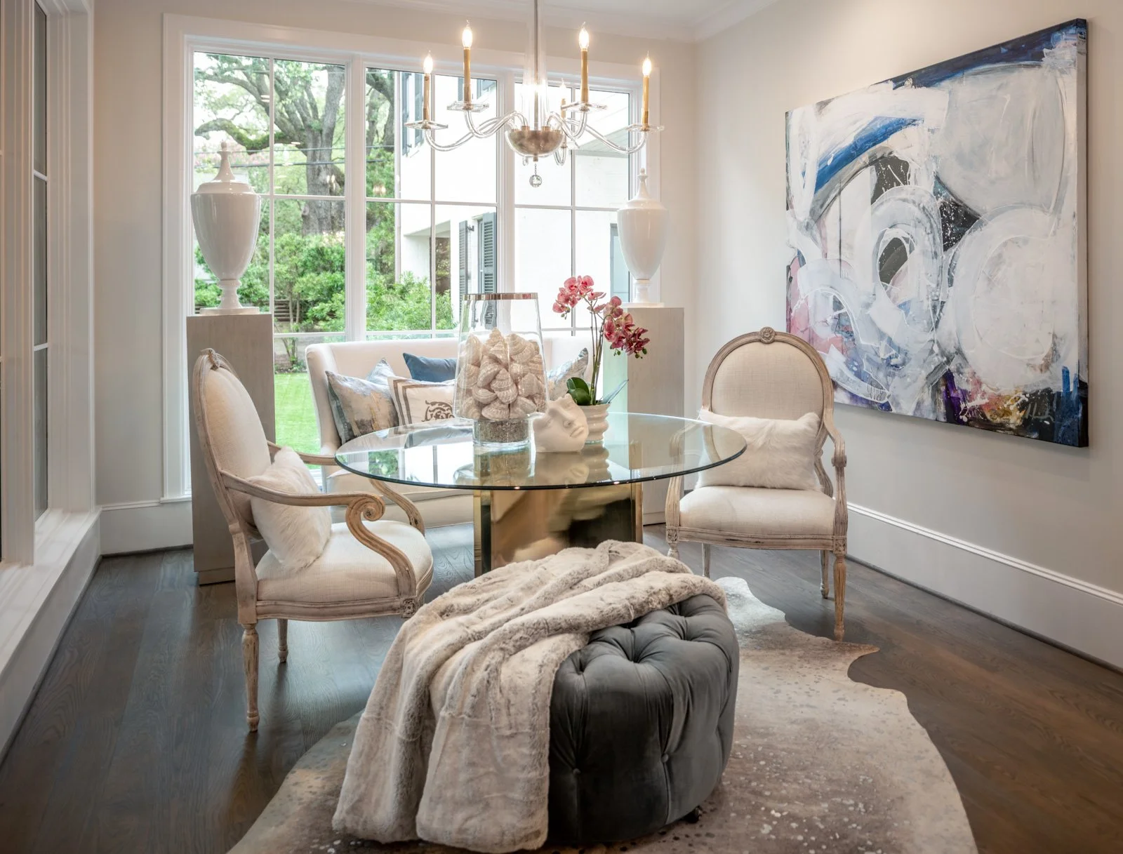 Luxury River Oaks dining room designed by The Design Firm in Houston, featuring a round glass dining table, elegant upholstered chairs, statement chandelier, large abstract artwork, and a tufted ottoman.