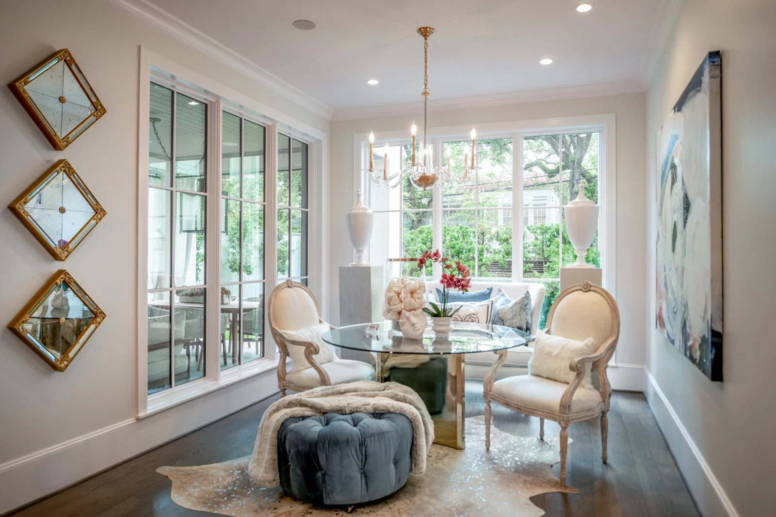 River Oaks dining room designed by The Design Firm in Houston, featuring a round glass dining table, upholstered chairs, elegant chandelier, tufted ottoman, decorative mirrors, and large windows overlooking the garden.