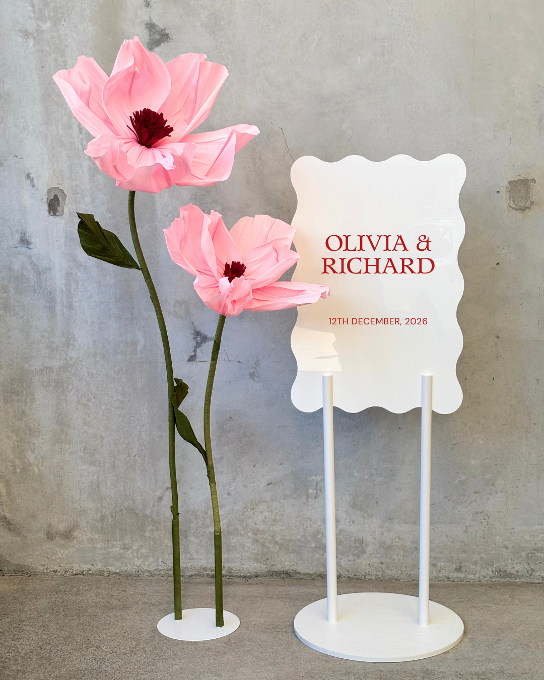 Decorative wedding sign with scalloped edges displaying names Olivia and Richard and the date December 12, 2026, positioned next to large pink paper flowers resembling poppies with green stems and leaves, against a textured concrete wall.