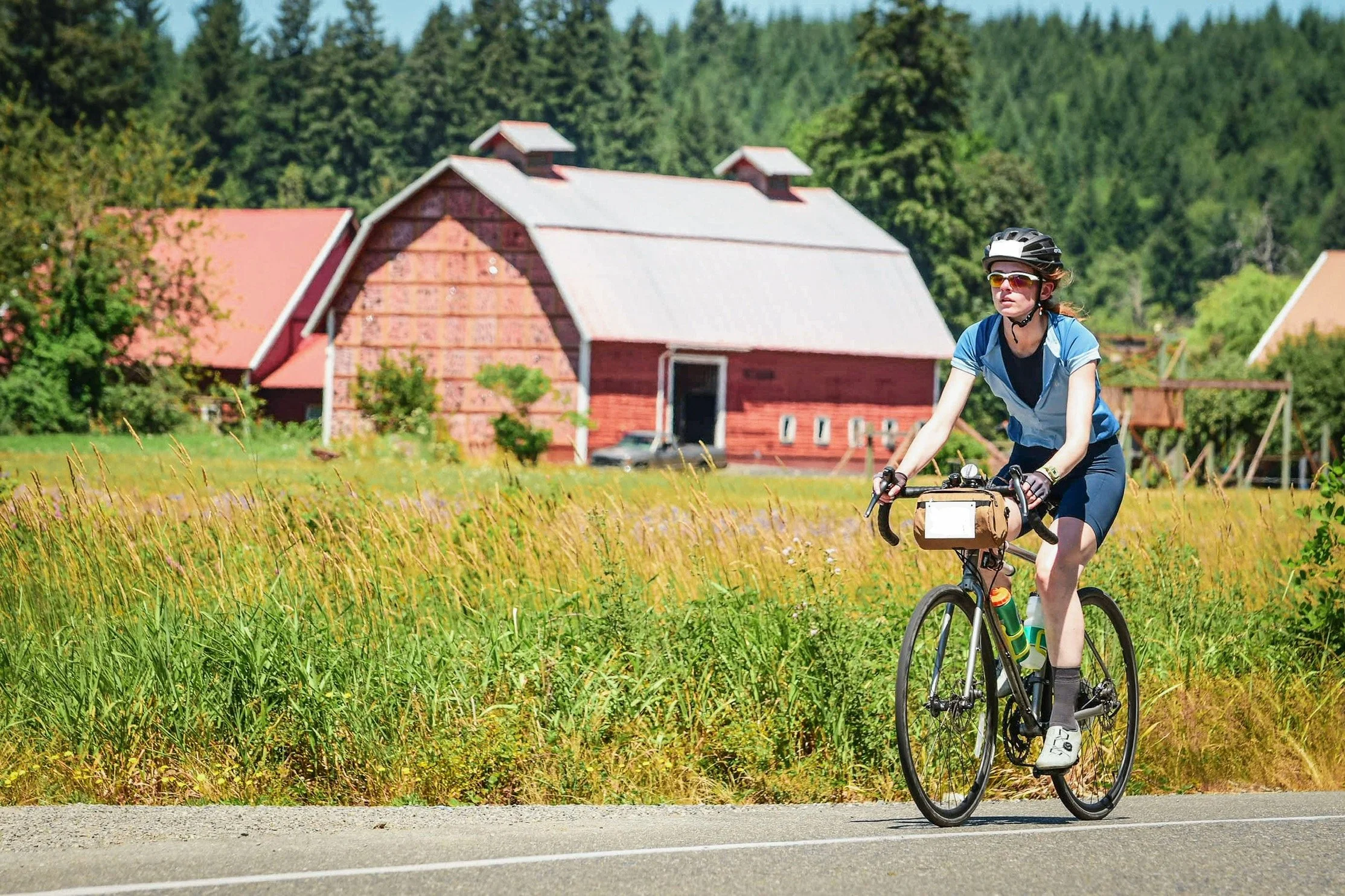 Woman cycling on a rural road with a red barn and green trees in the background.