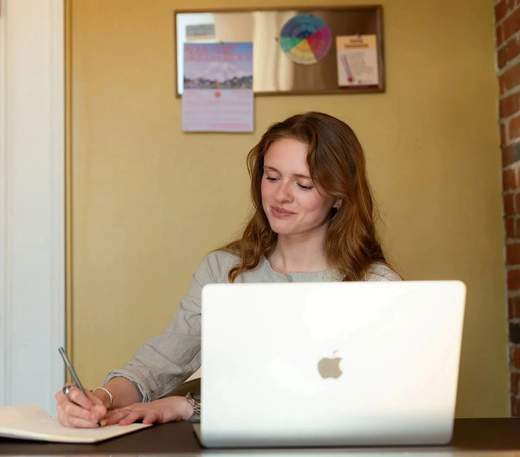 A young woman with red hair, wearing a beige top, sitting at a desk with a silver pen writing in a notebook. She is in front of an open laptop and appears to be focused. The workspace has a yellow wall with a framed pinwheel and a calendar on it, and a brick wall to the side.