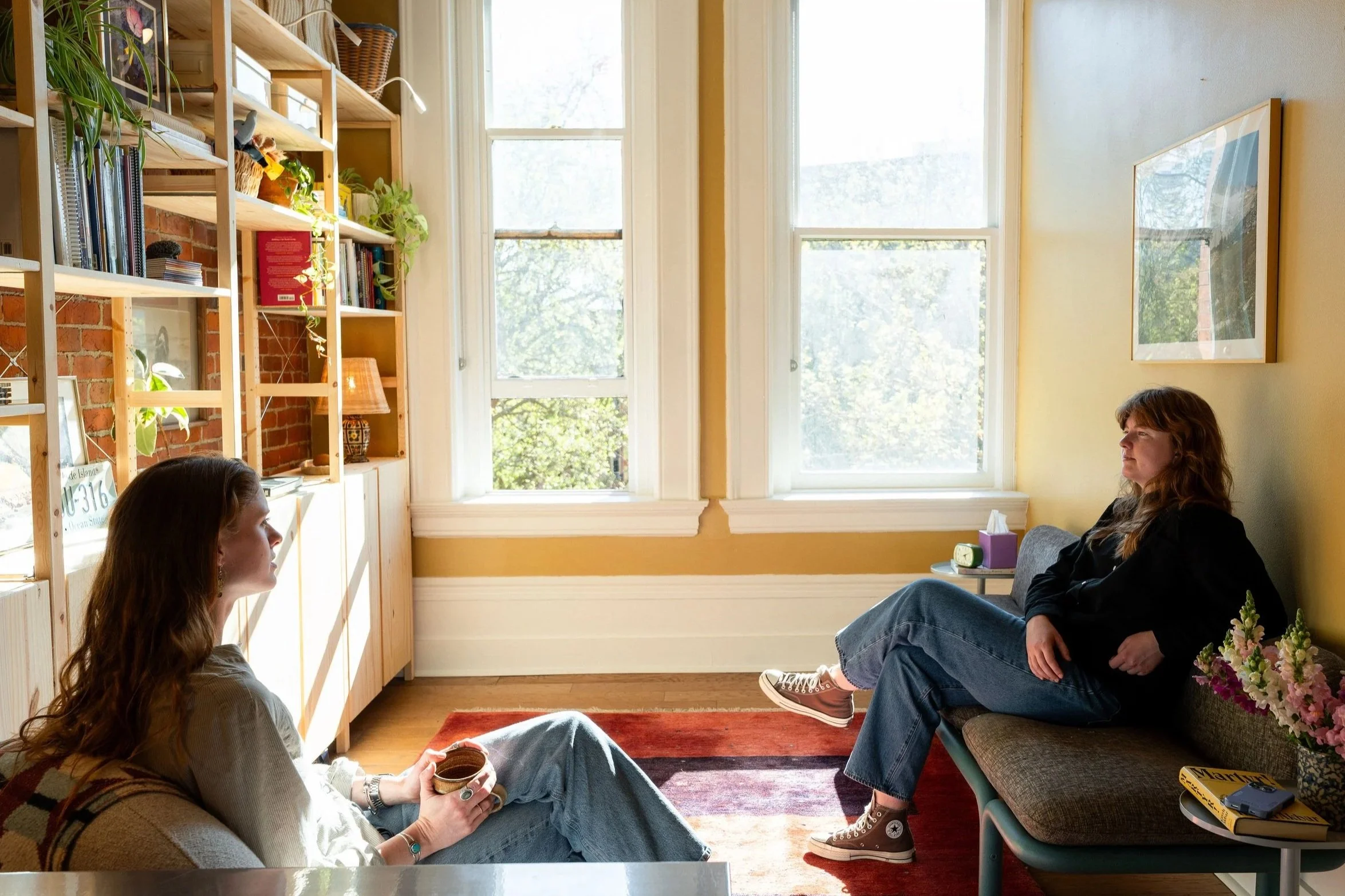A cozy living room with large windows letting in natural light. Two women with long hair are sitting on the floor and on a couch, engaging in conversation. A wooden bookshelf filled with books and plants is against the brick wall. A small side table with a lamp is next to the woman on the couch.