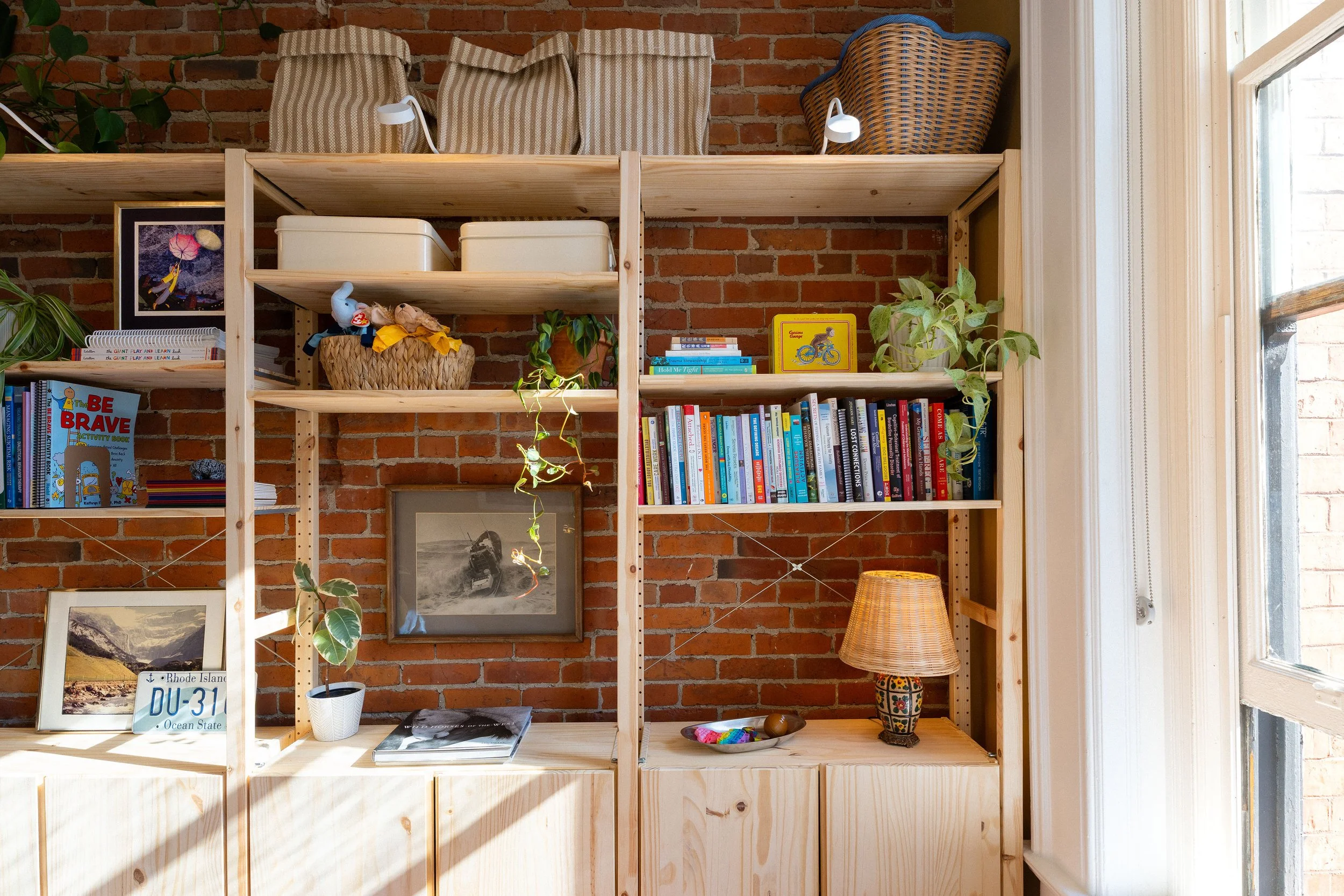 A wooden bookshelf with multiple shelves against a brick wall filled with books, decorative items, and plants, near a window with natural light.