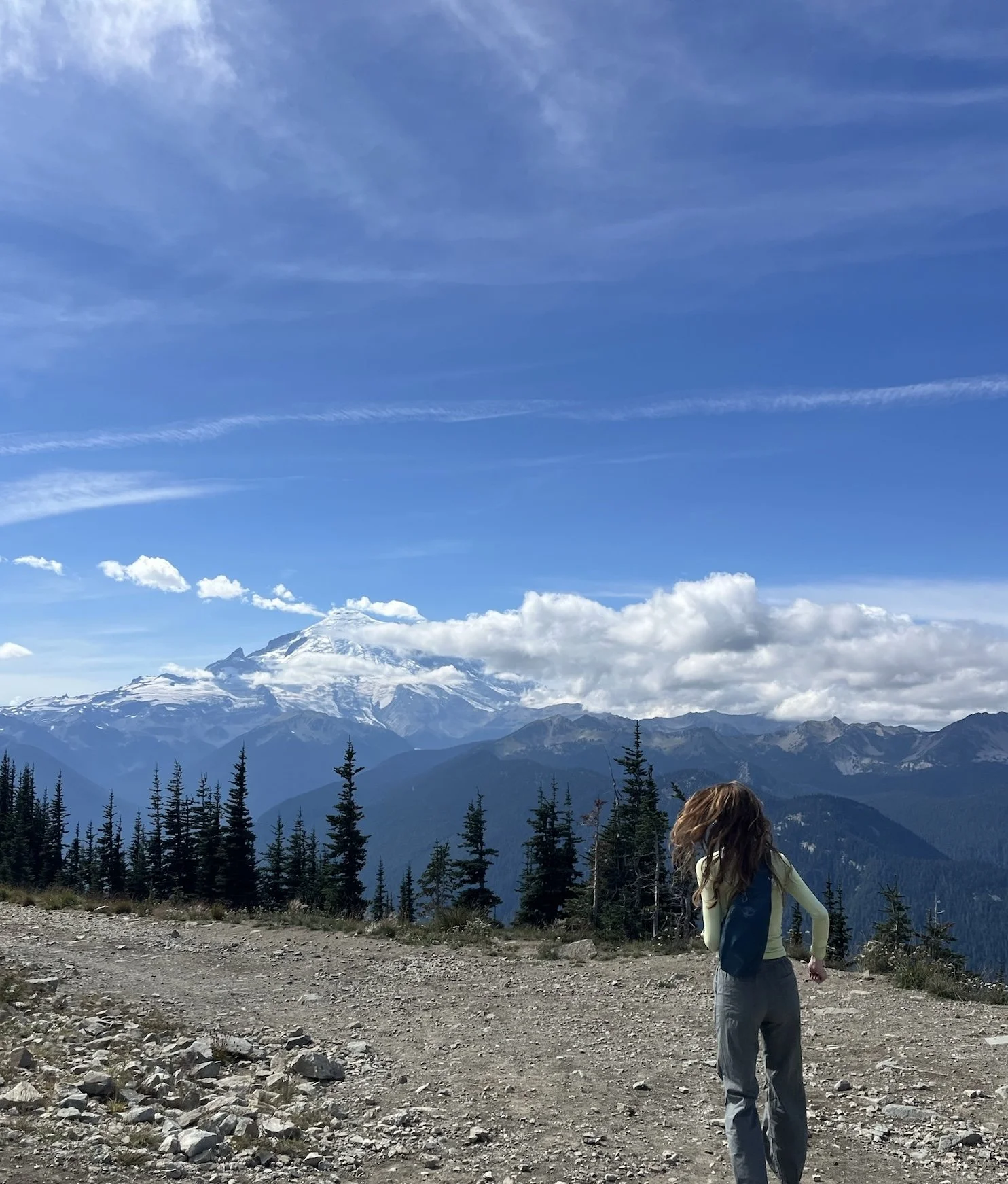 A woman with long hair wearing a yellow and blue jacket and gray pants hiking on a rocky trail in a mountainous area with snow-capped peaks, evergreen trees, and a partly cloudy sky in the background.