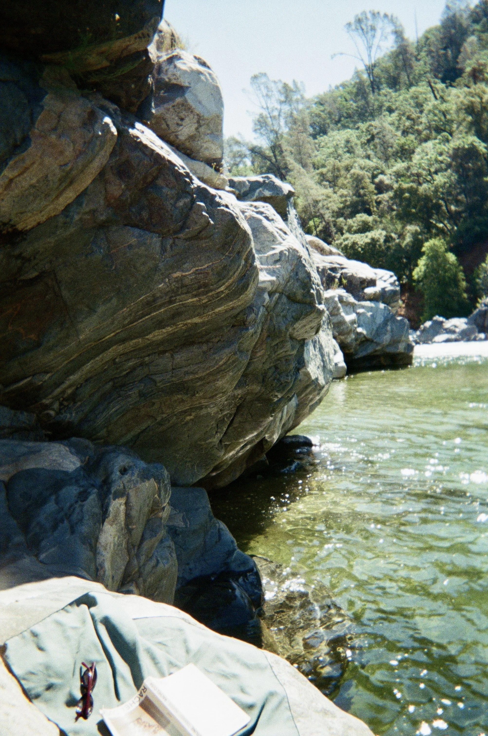 Large rock formation along a riverbank with trees in the background and a book and glasses resting on a cloth in the foreground.
