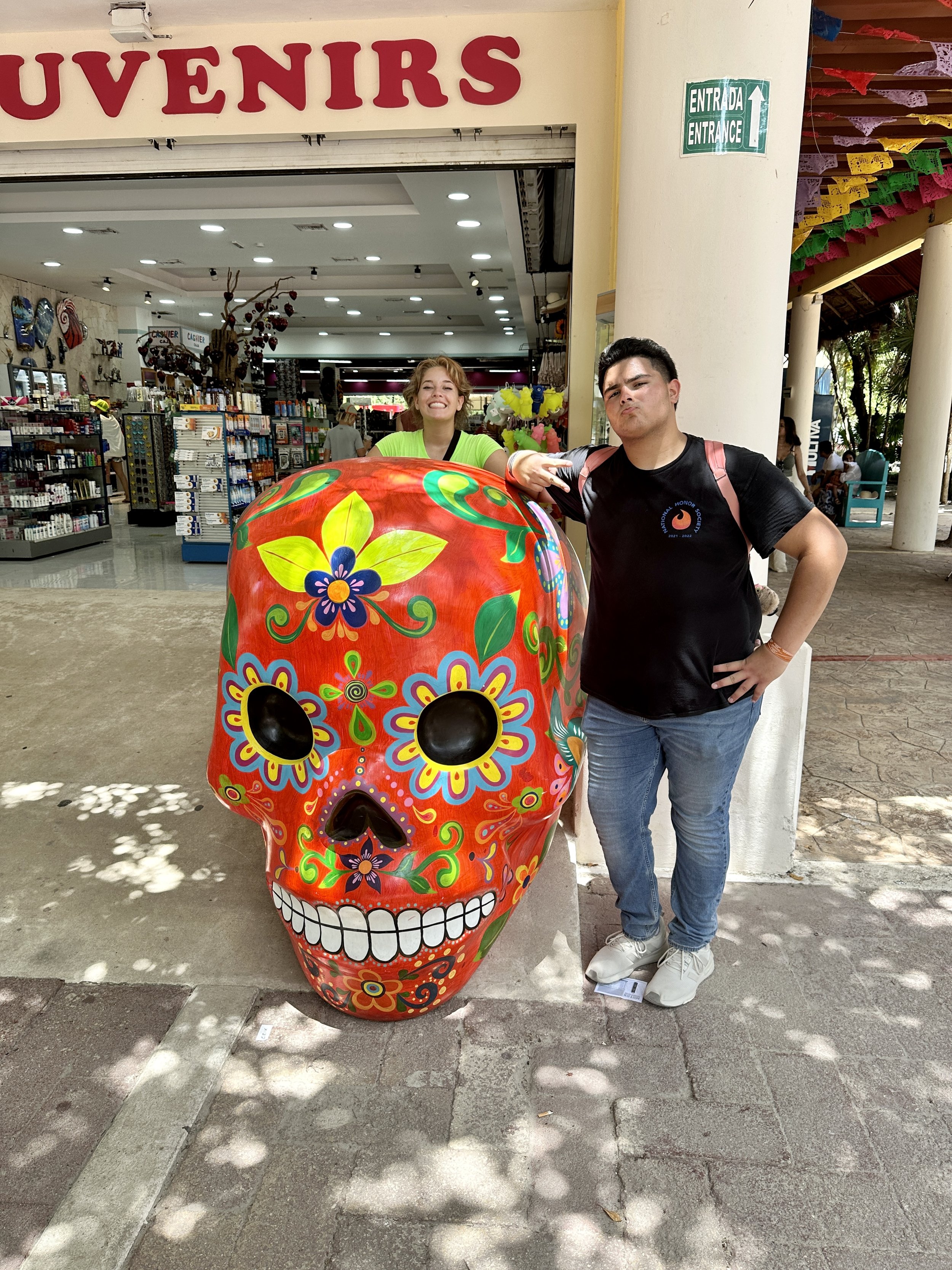 Two people posing with a large decorative Day of the Dead sugar skull in front of a souvenir shop entrance, with colorful papel picado banners above.
