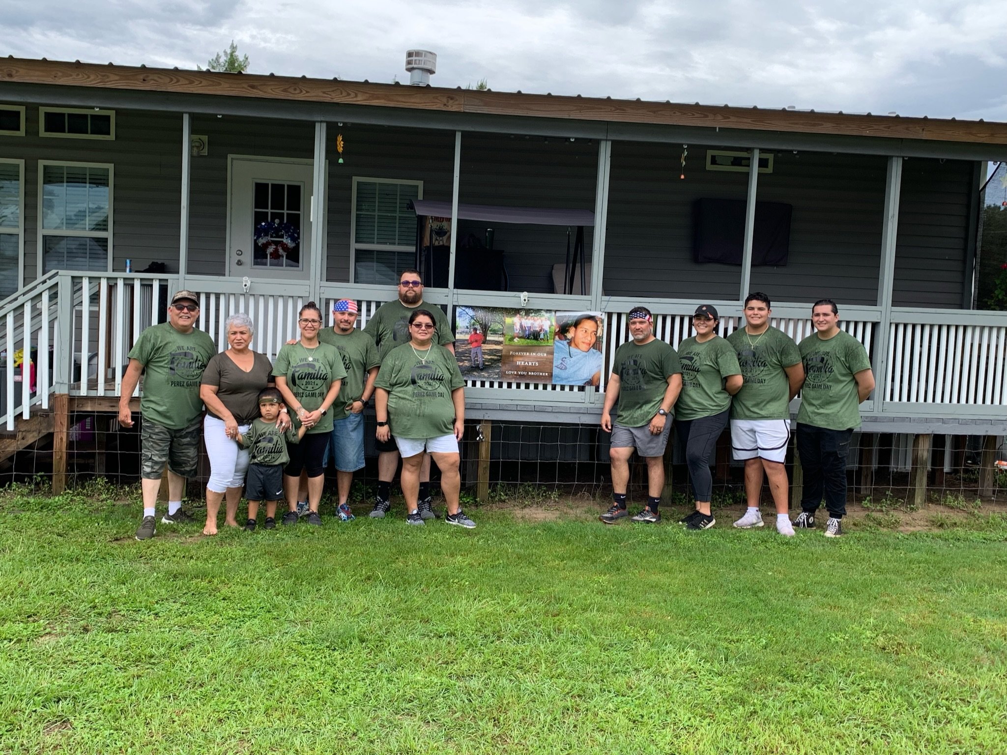 Group of people in matching green shirts posing in front of a house with a porch. They are standing on grass, and there is a banner with a portrait attached to the porch.
