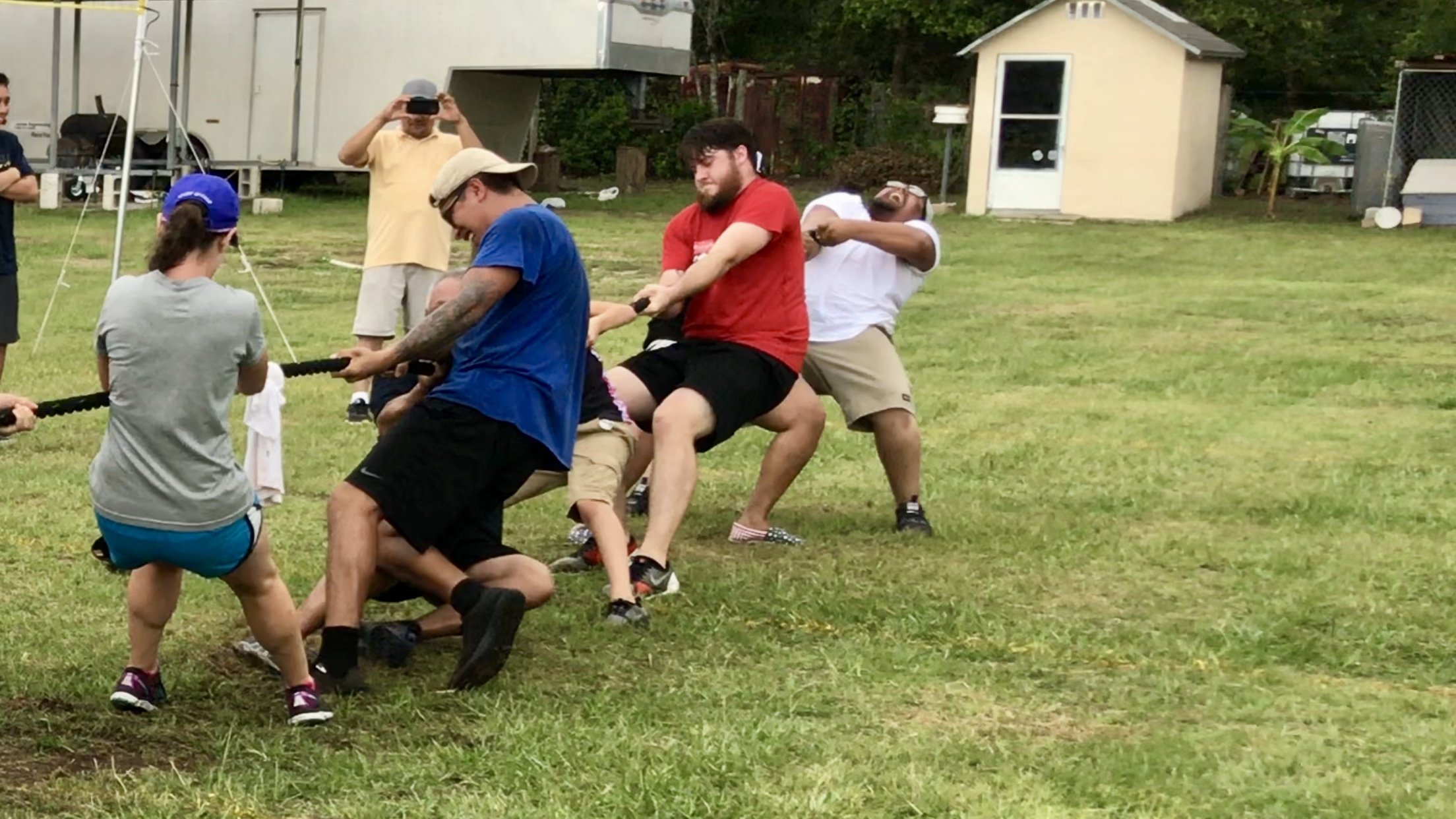 Group of people playing tug-of-war on grass, with some pulling a rope and others observing.