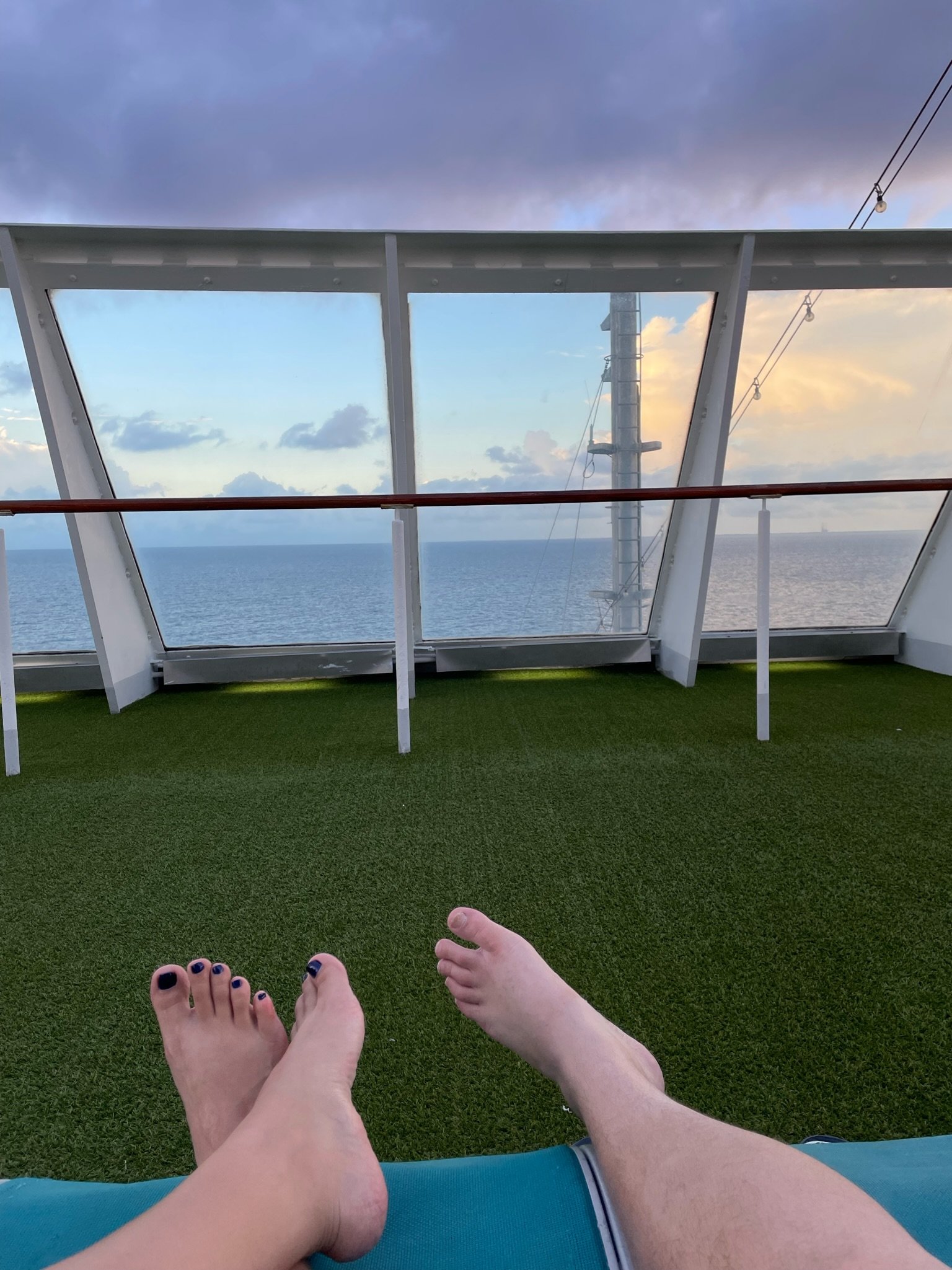Two people relaxing on a cruise ship deck, with feet visible, overlooking the ocean and sky.