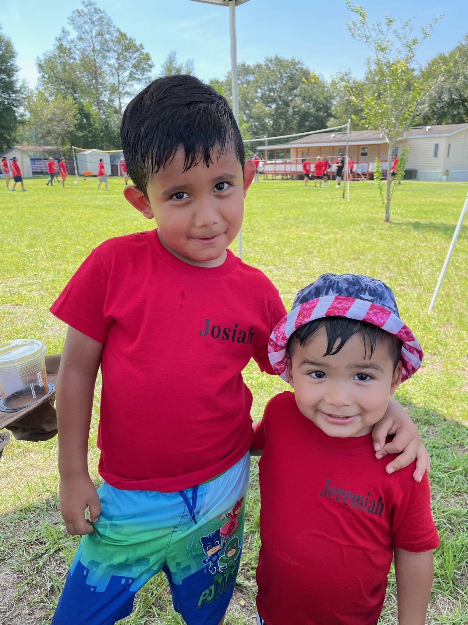 Two young children wearing red shirts, smiling outdoors on a sunny day. The older child has 'Josiah' on their shirt and the younger has 'Jeremiah' on theirs. They are standing on grass with a sports activity happening in the background.