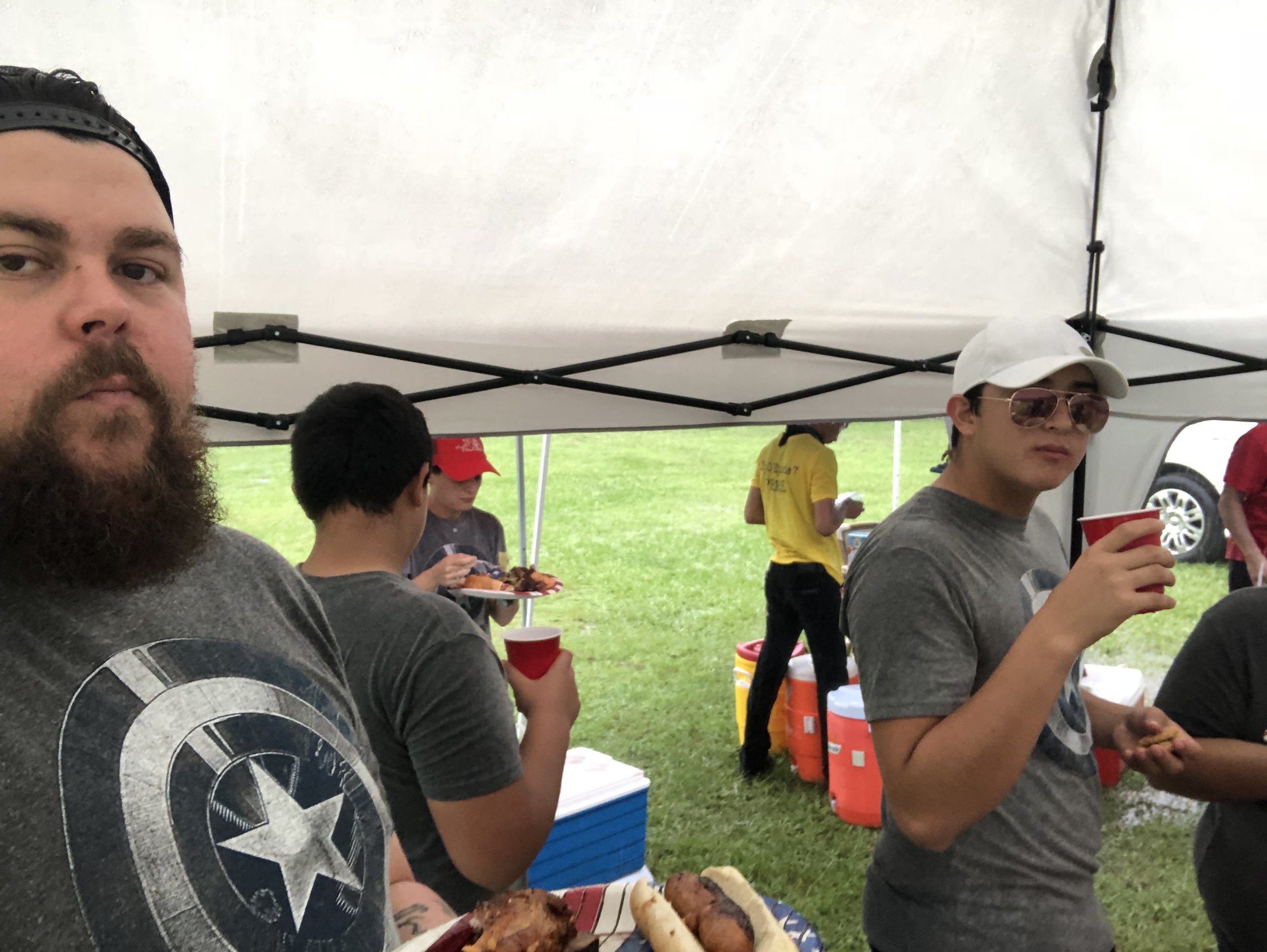 Group of people under a tent at an outdoor gathering, holding food and drinks. Some wear casual clothing including a t-shirt with a star emblem, and one individual wears sunglasses and a white cap. A grassy area and beverage coolers are visible in th