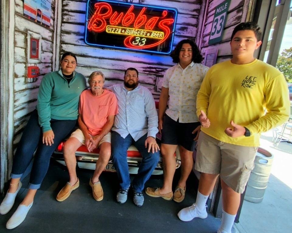 Five people posing in front of a neon sign reading 'Bubba's Pizza, Burgers & Beer' and '33'.