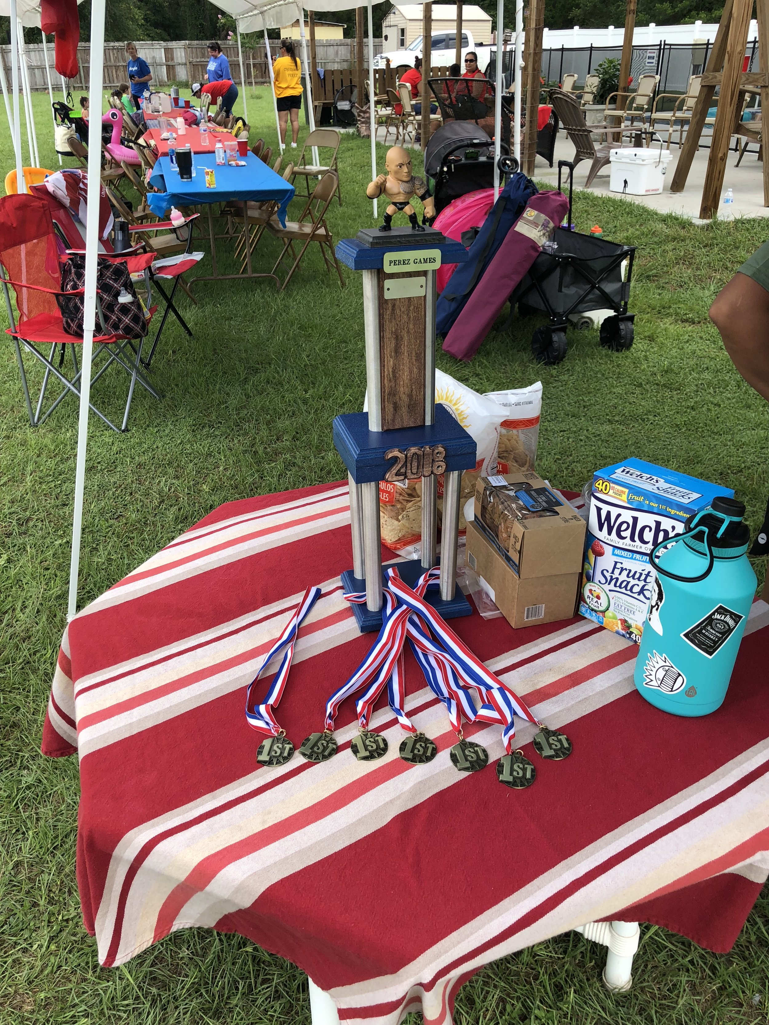 Outdoor event setup with a table holding a 2018 trophy labeled "Perez Games," several first-place medals, and snack items. Nearby are chairs, a water bottle, and a canopy with more tables and people in the background.