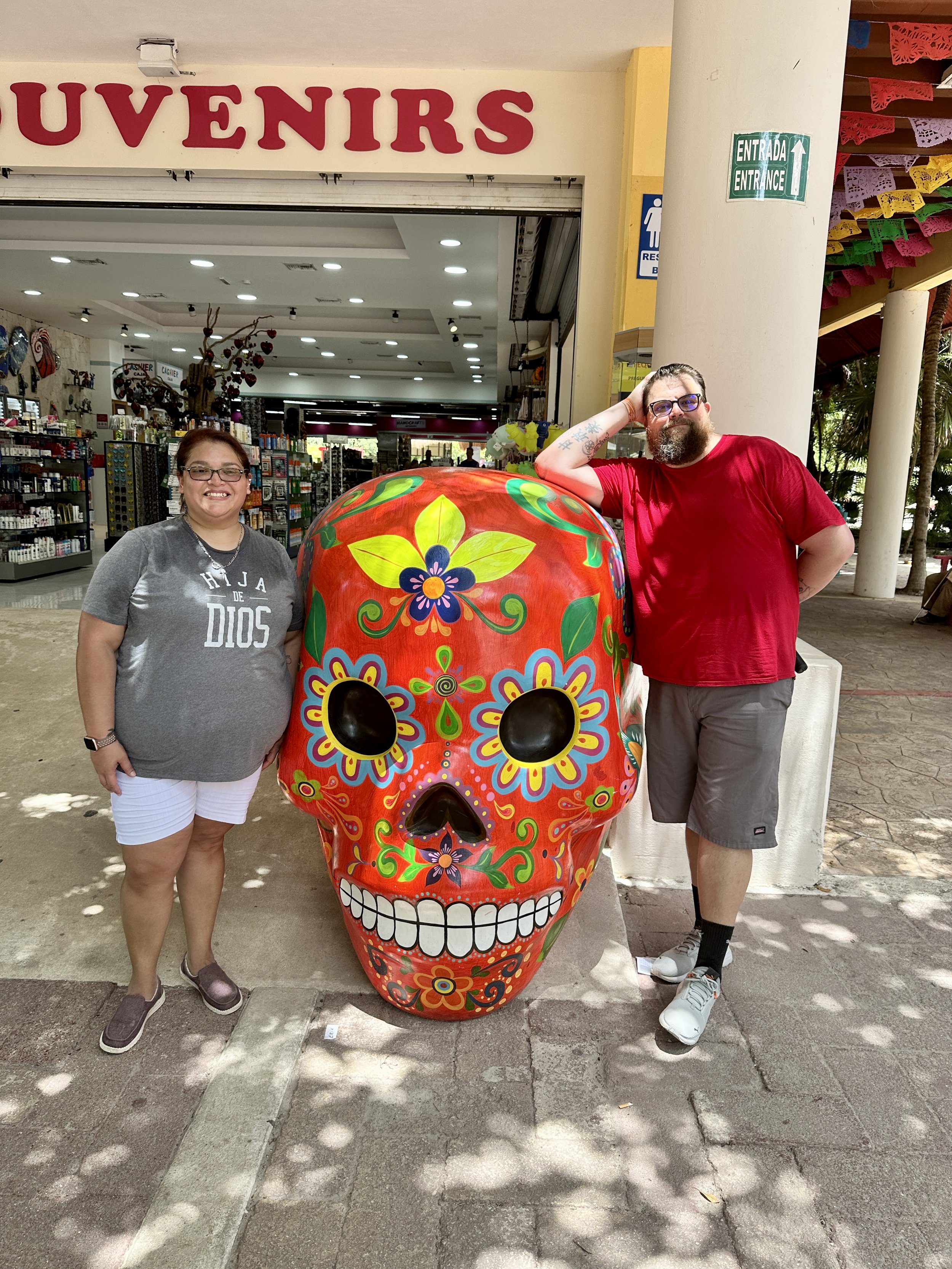 Two people standing beside a large, colorful Day of the Dead skull decoration outside a souvenir shop. The skull features vibrant floral patterns.