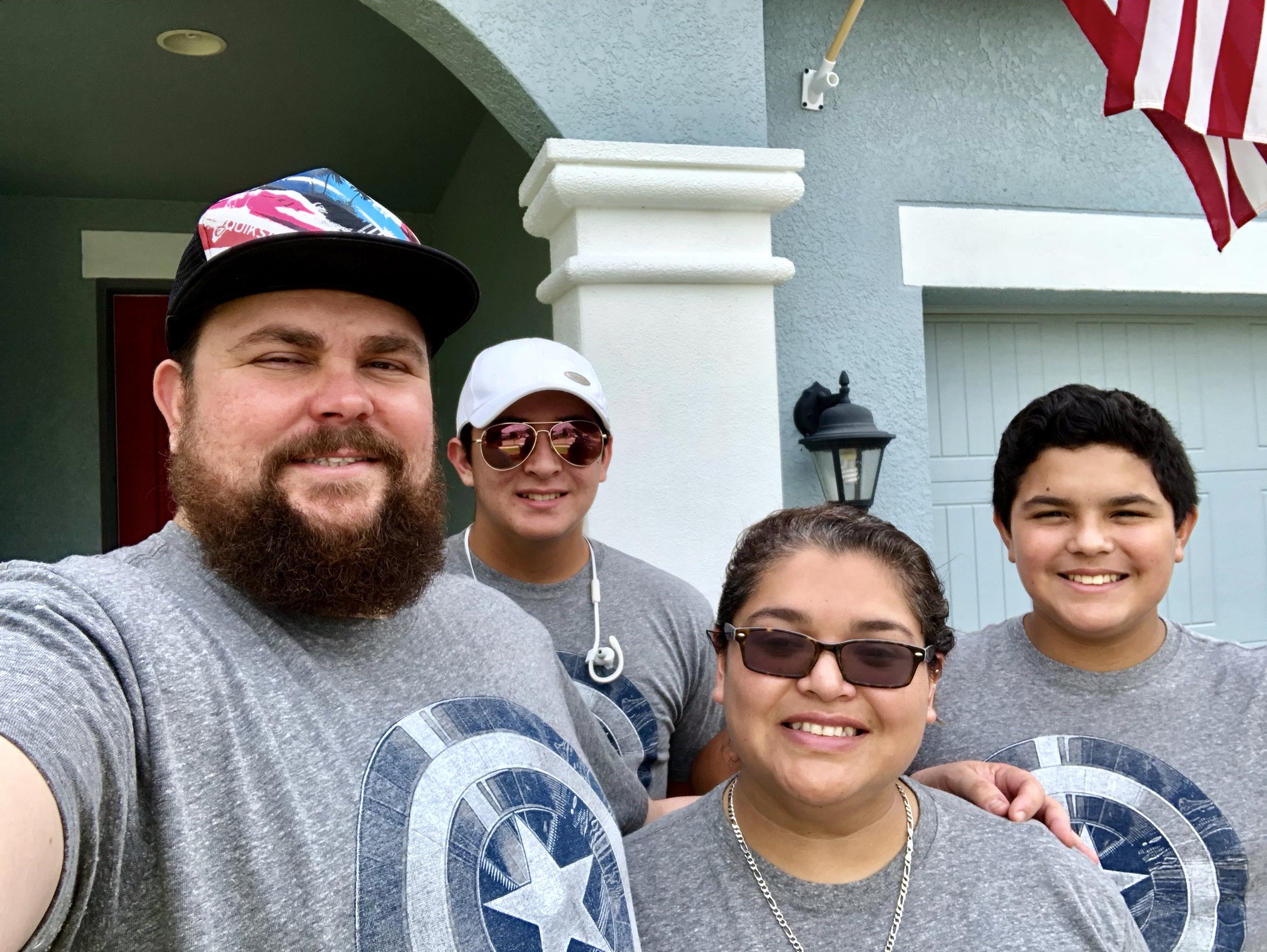 Family of four wearing matching gray T-shirts with a Captain America shield. They are standing outdoors in front of a house with a visible American flag.