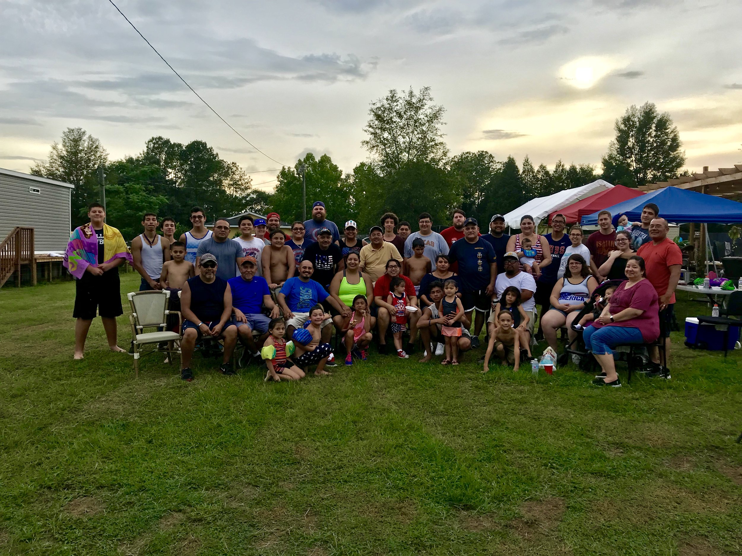 Large group of people gathered outdoors on grass, with tents in the background, during a cloudy day.