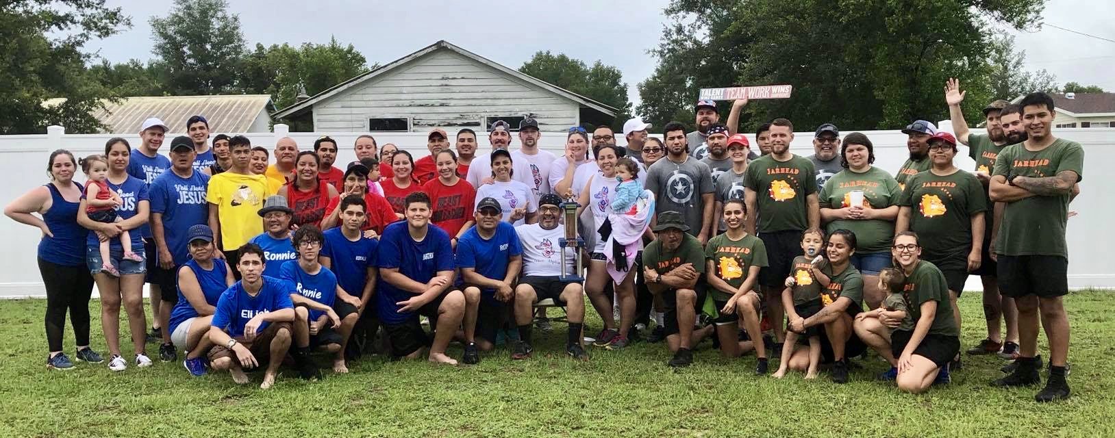A group of diverse people standing and sitting on a grassy field in front of a house, wearing different themed T-shirts in blue, red, yellow, gray, and green. Some are holding children or signs. A white fence and trees are visible in the background.