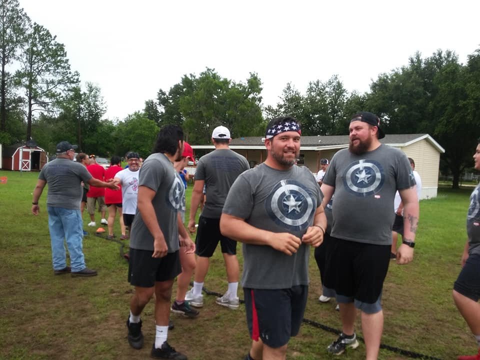 Group of men wearing matching Captain America t-shirts outdoors, some shaking hands, in a grassy area with trees and a building in the background.