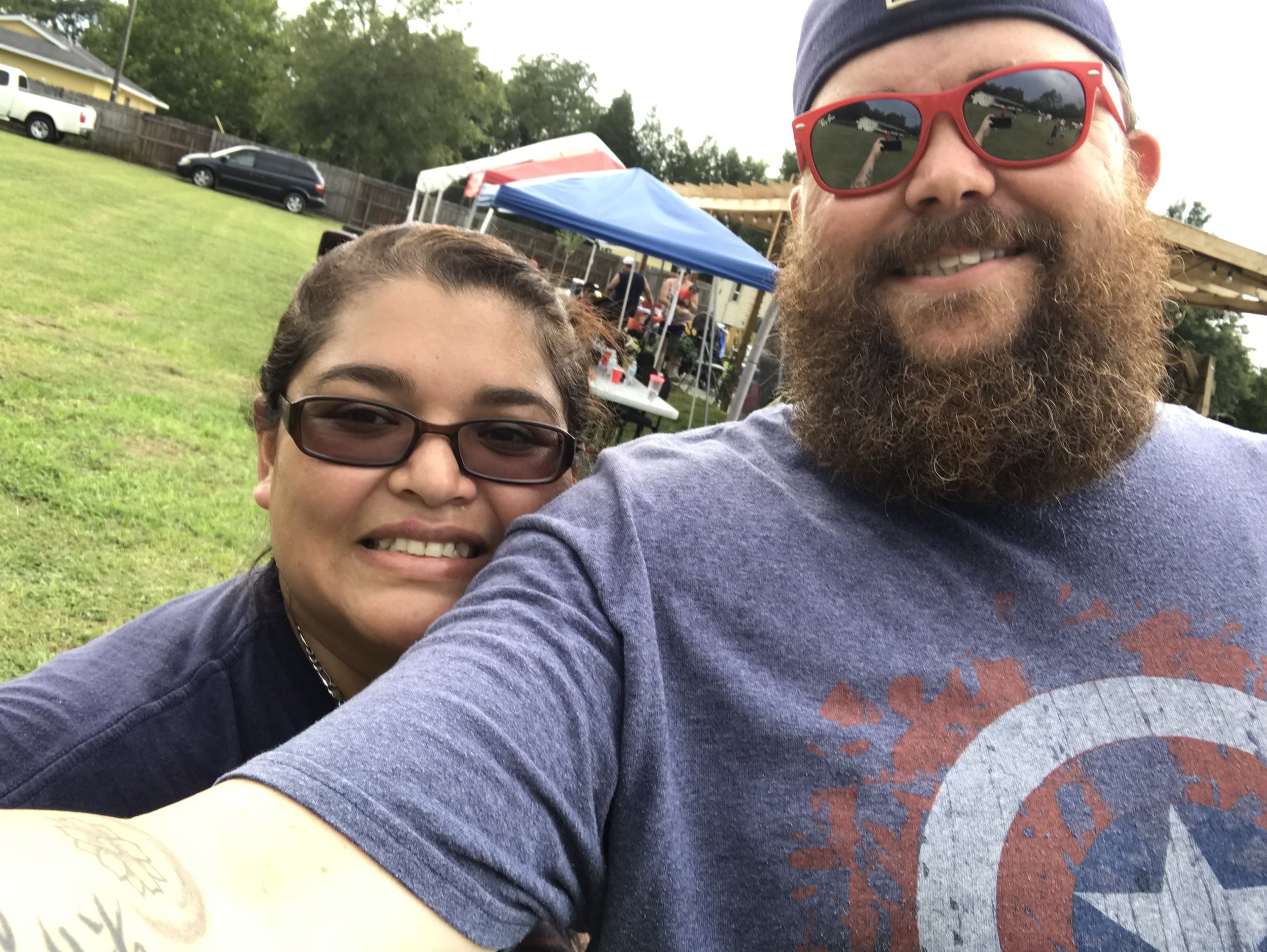 A person wearing sunglasses and a beard smiles next to another person wearing glasses, in an outdoor setting with tents and grass.