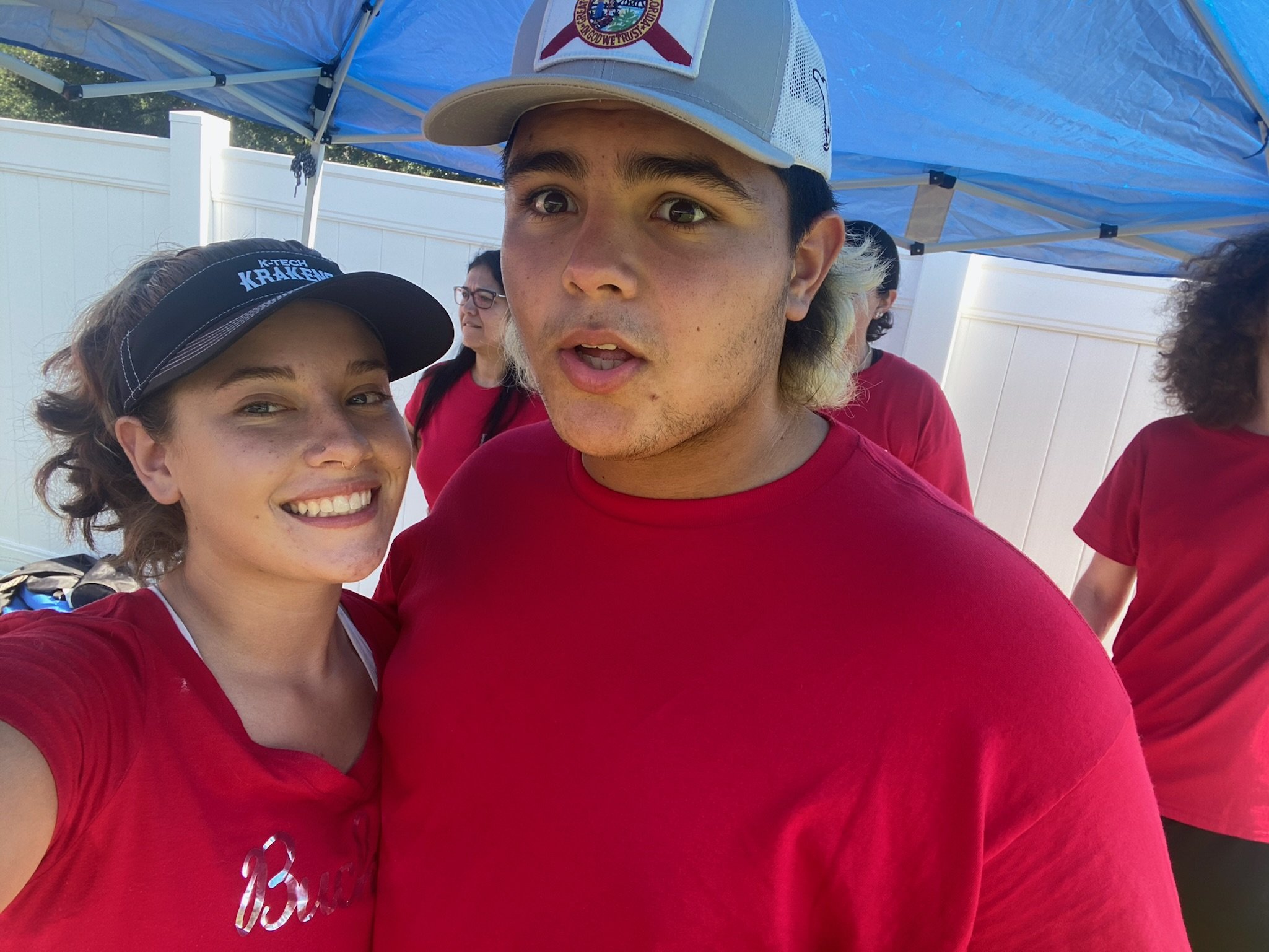 Two people in red shirts under a blue canopy, one smiling, the other with a surprised expression. A woman wearing a visor and a man in a cap are visible, with a group of people in the background.