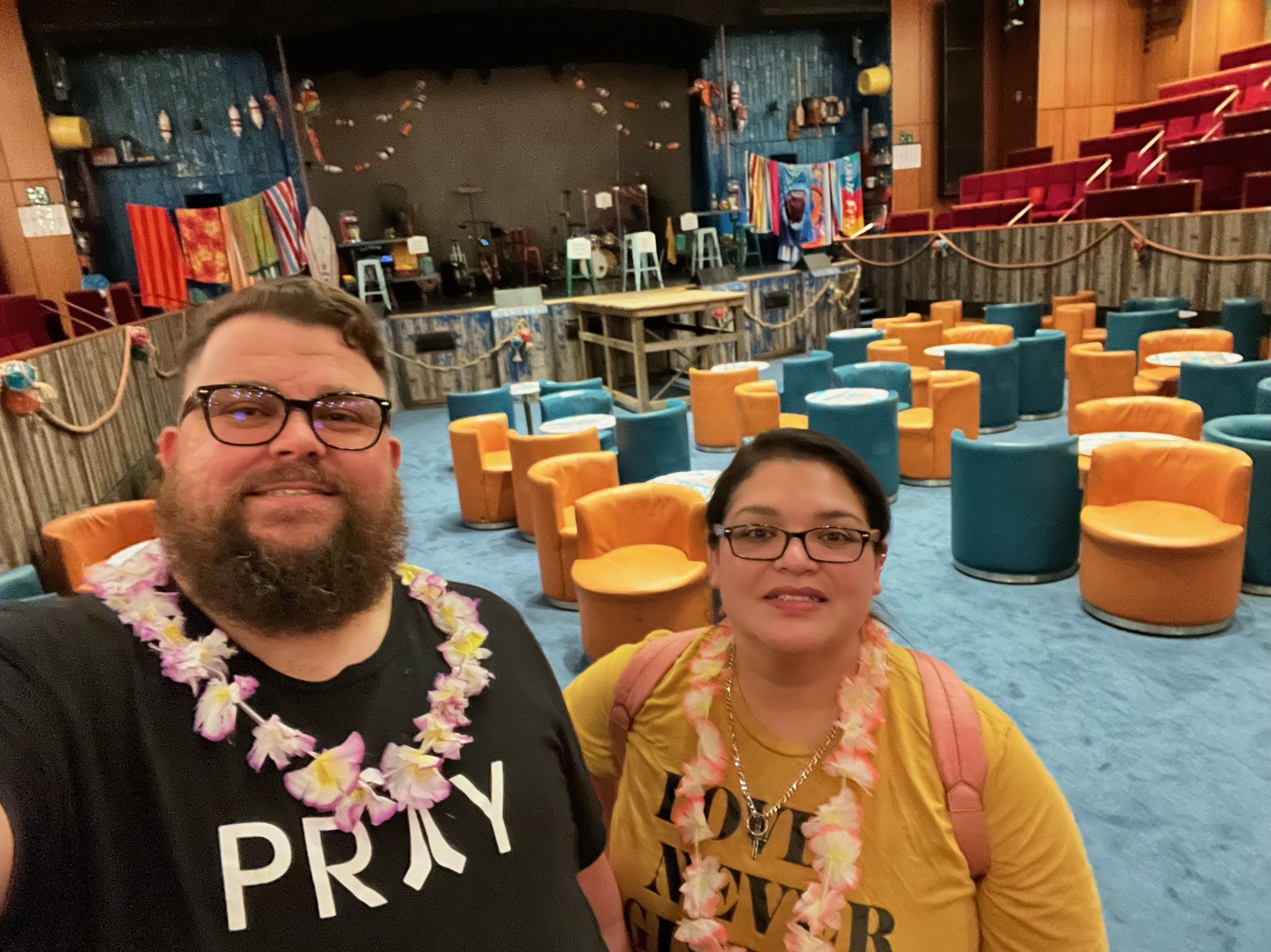 Two people wearing leis inside a colorful, relaxed lounge with blue and orange seating, a stage, and nautical-themed decorations.