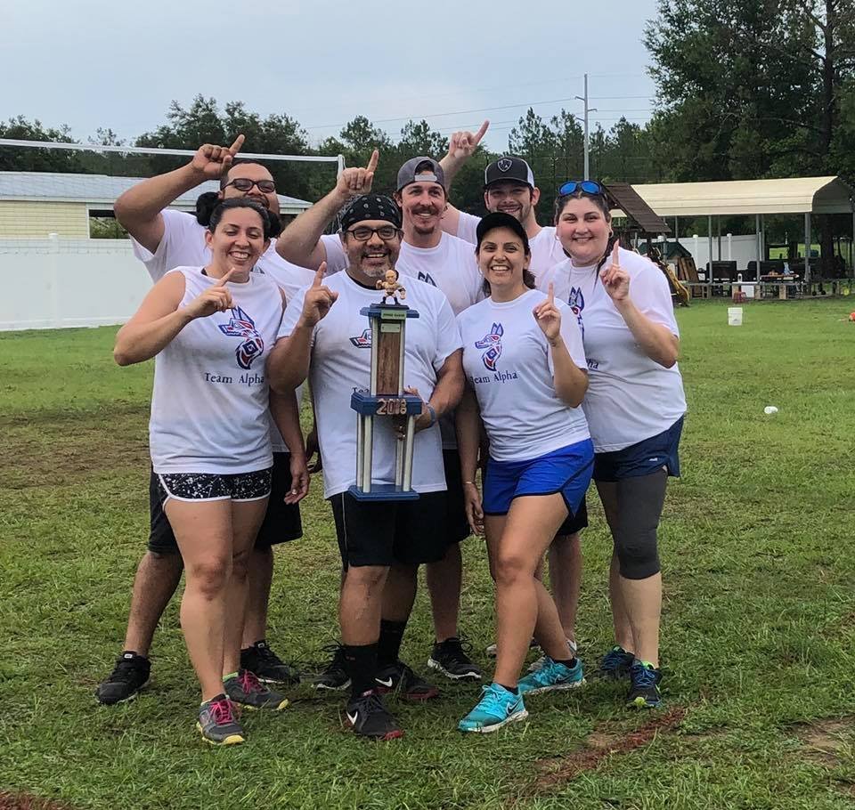 Group of seven people outdoors wearing matching "Team Alpha" shirts, holding a trophy and showing number one gestures. They are standing on a grassy field with a white fence and trees in the background.