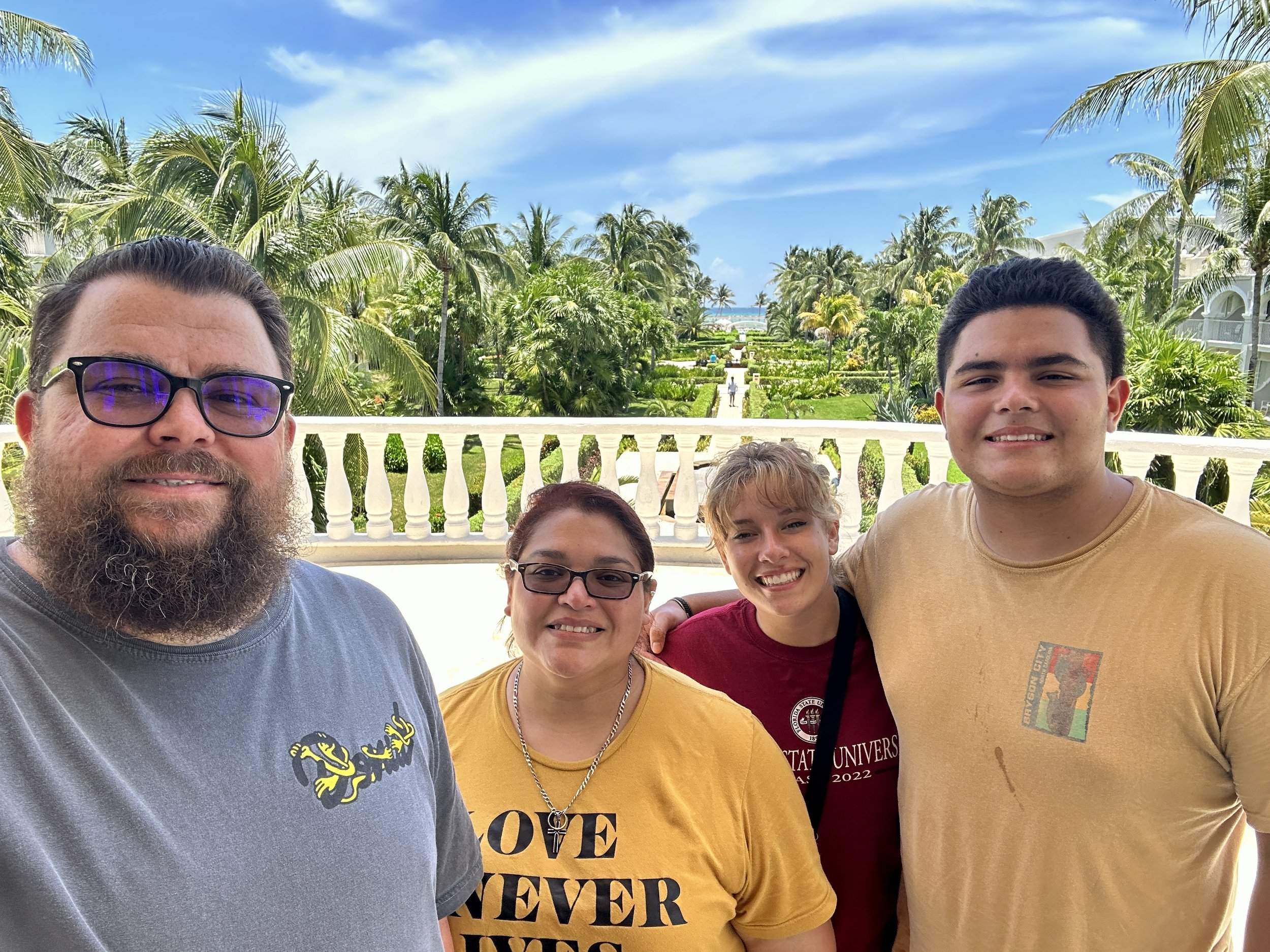 A group of four people smiling on a balcony with tropical greenery and palm trees in the background. They appear to be enjoying a sunny day.