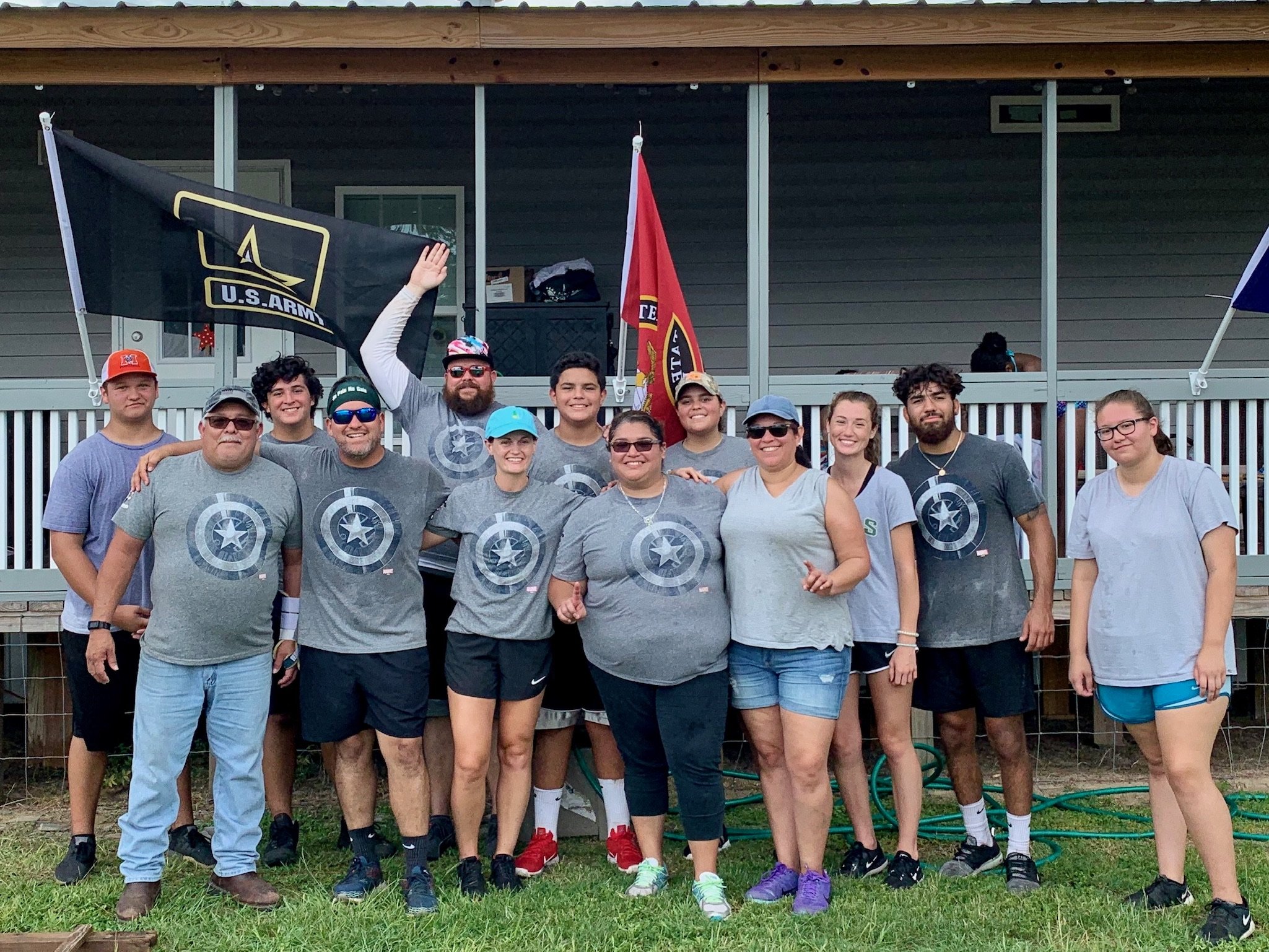 Group of people posing outdoors in matching t-shirts, with U.S. Army and Texas flags in the background.