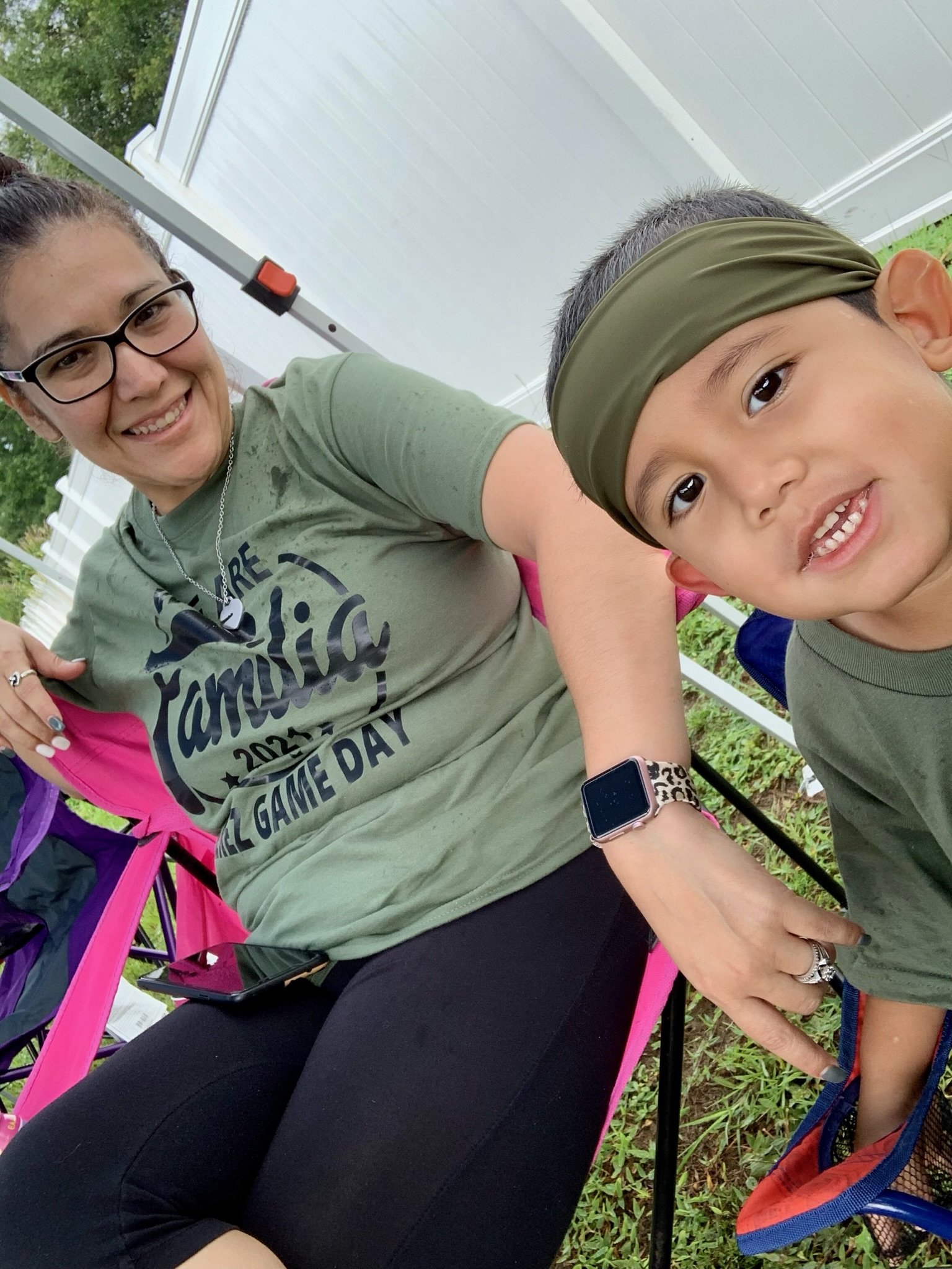 A woman and a young child sitting outdoors on foldable chairs, both wearing green shirts. The woman is smiling and wearing glasses and a smart watch. The child is also smiling and wearing a headband. The background includes a white fence and some gre