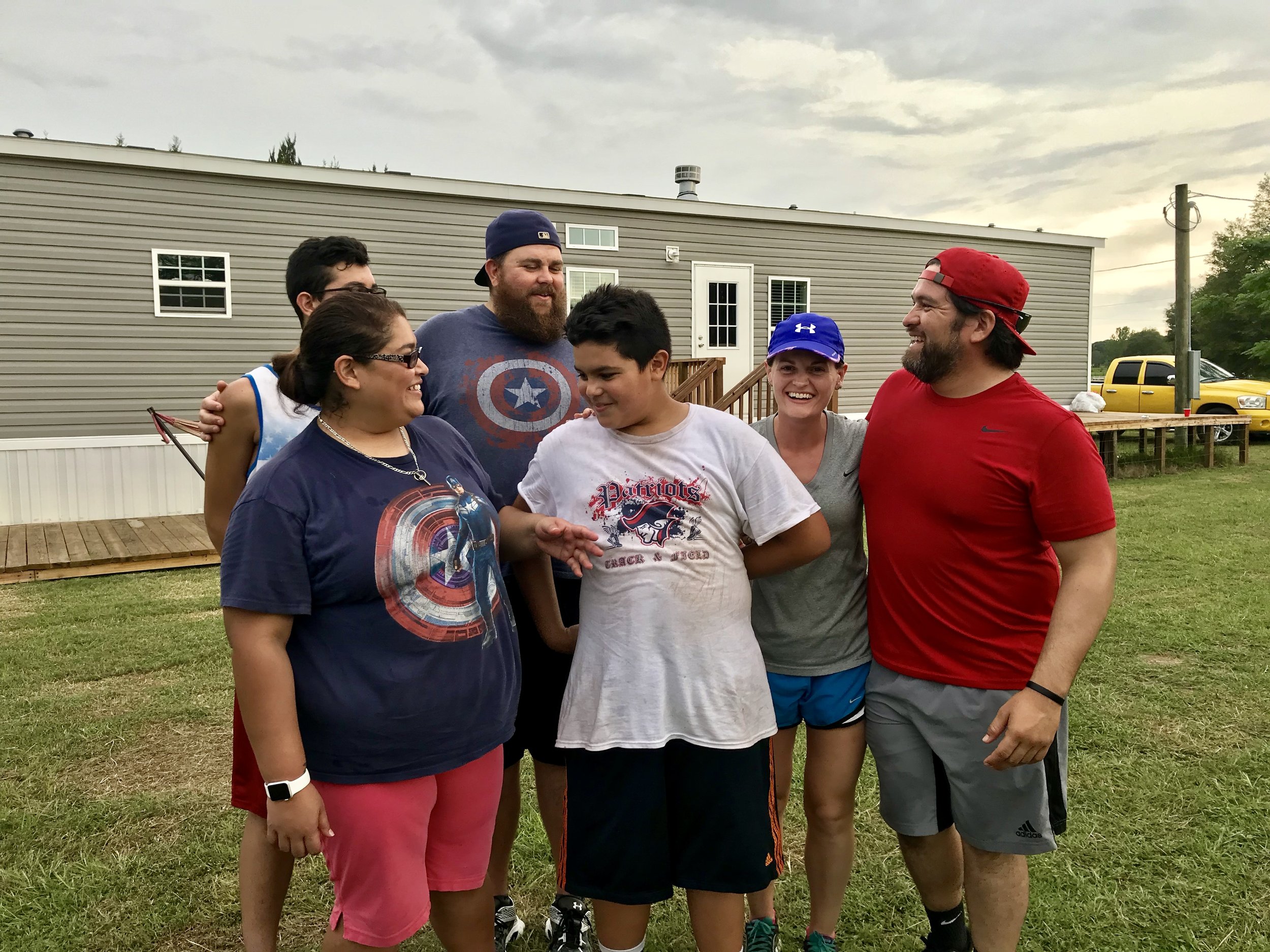 A group of six people standing together outdoors, smiling and laughing. They are casually dressed, and there is a mobile home and a yellow truck in the background. The group appears to be enjoying a casual moment, possibly during a gathering or event