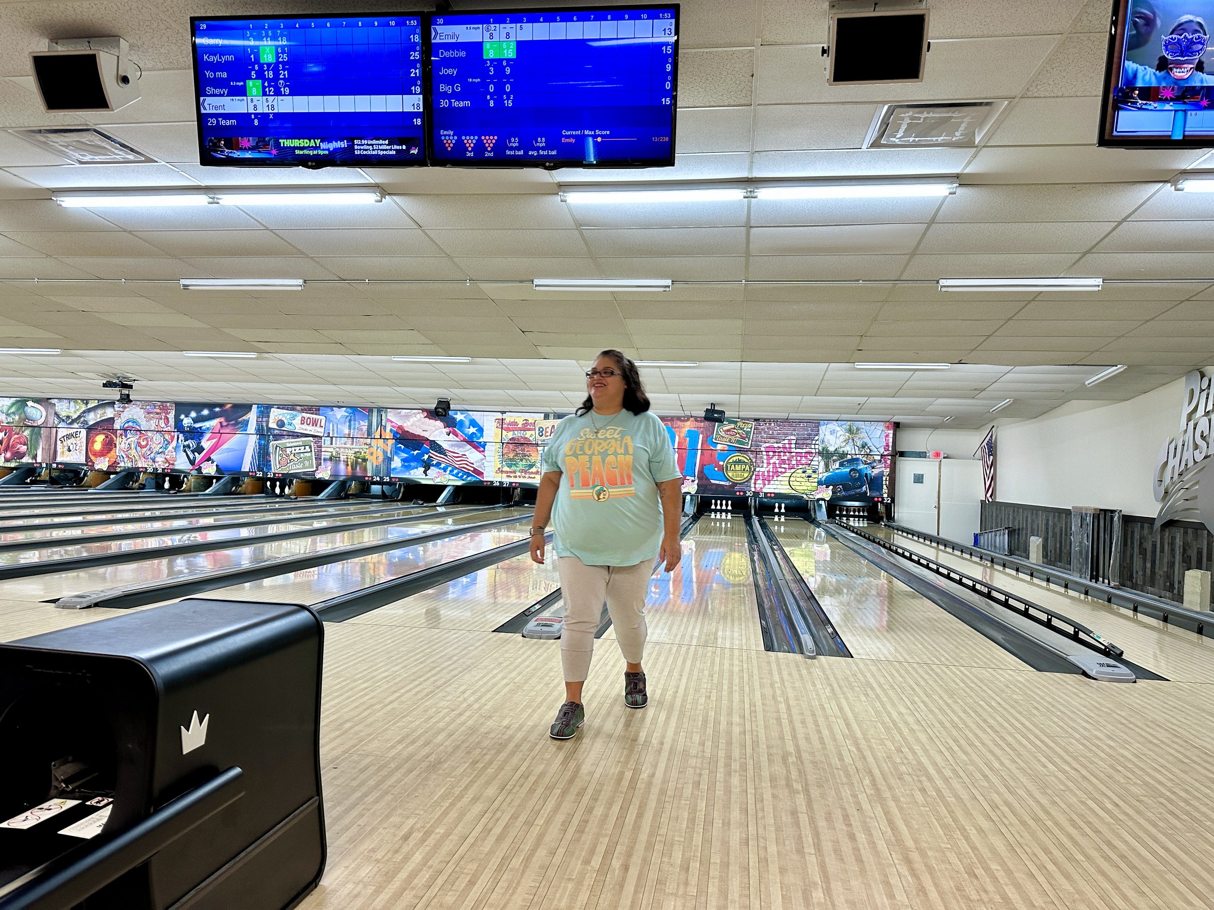 Person walking on a bowling alley lane with bowling scores displayed on screens above.