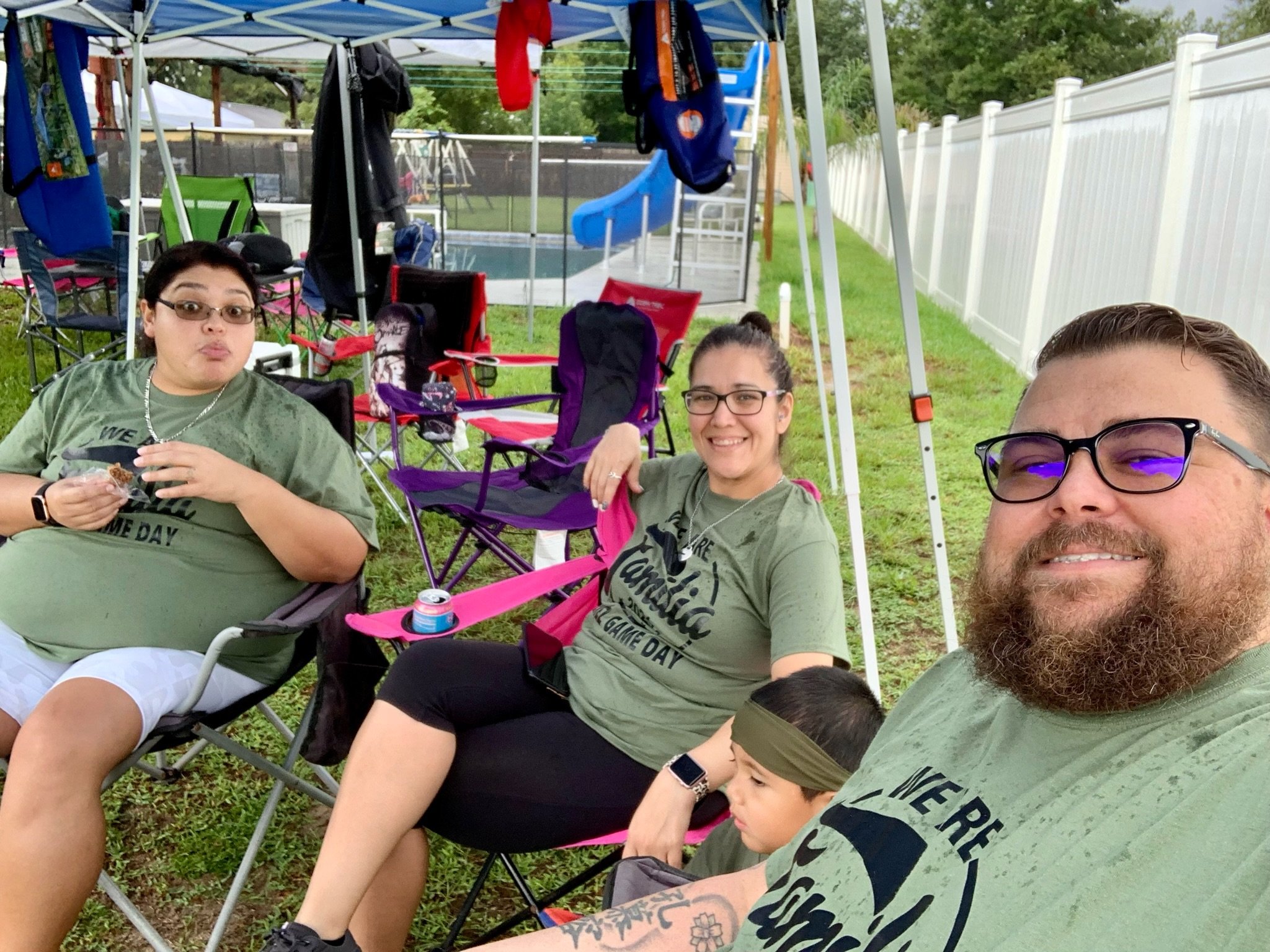 A group of people sitting outside under a canopy tent, wearing matching green shirts. They are seated on folding chairs on a grassy area next to a white fence. There are drinks and items around them, suggesting a casual gathering or event.