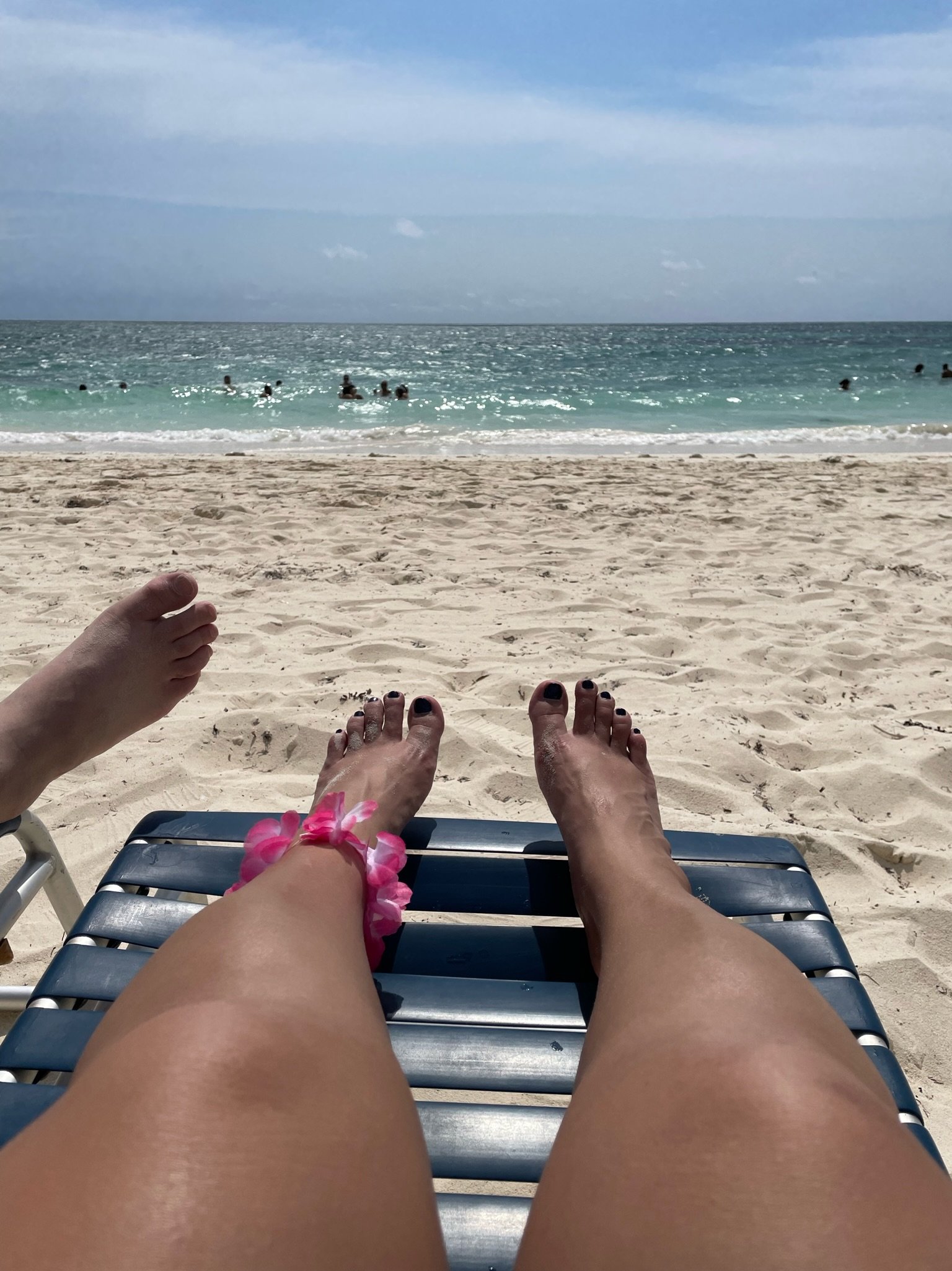 Person relaxing on a sun lounger at a sandy beach with ocean view and partly cloudy sky. Feet visible with pink floral bracelet.