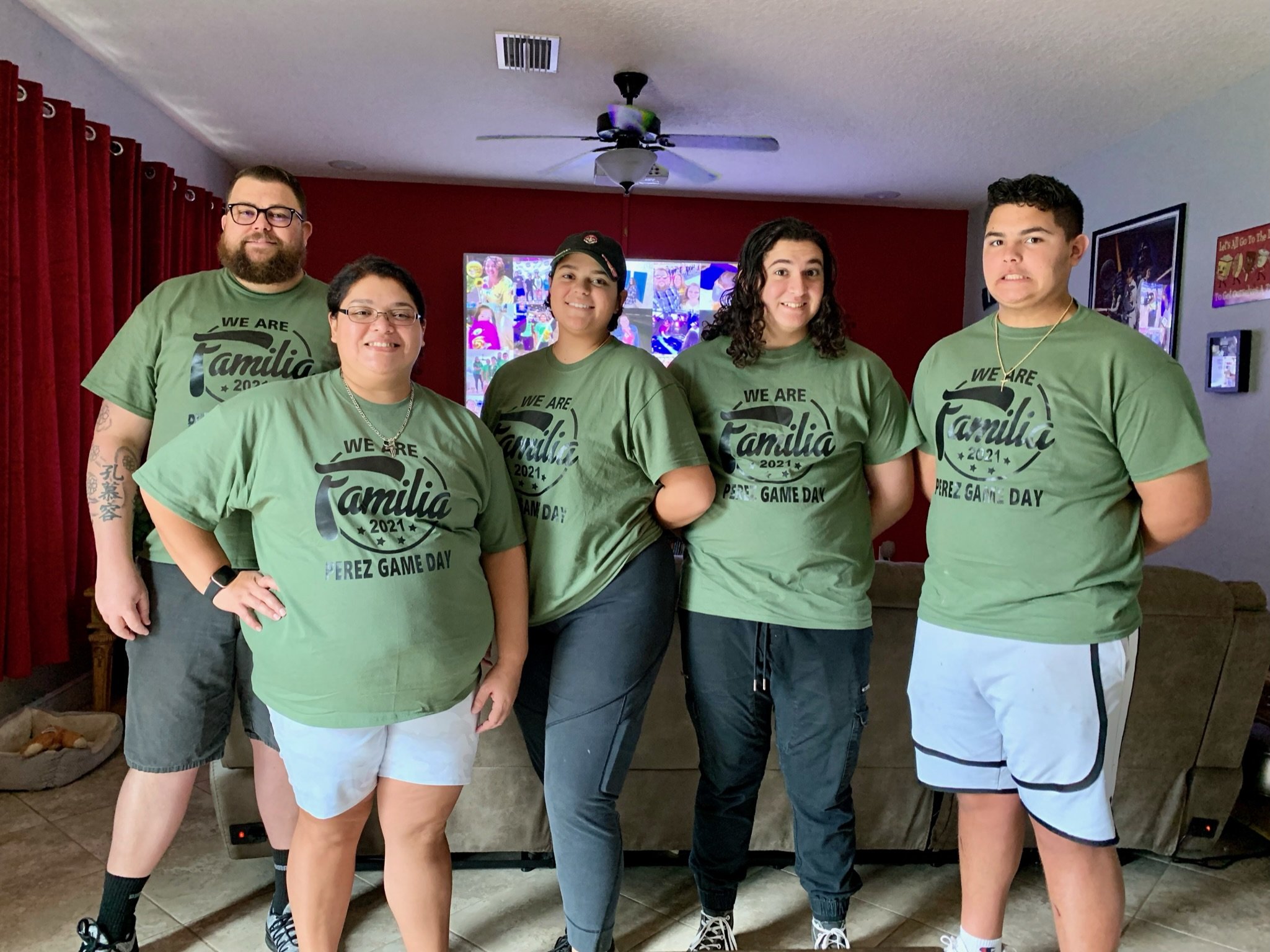 Group of five people wearing matching green t-shirts that read "We Are Familia 2021 Perez Game Day" standing in a living room with a television in the background.