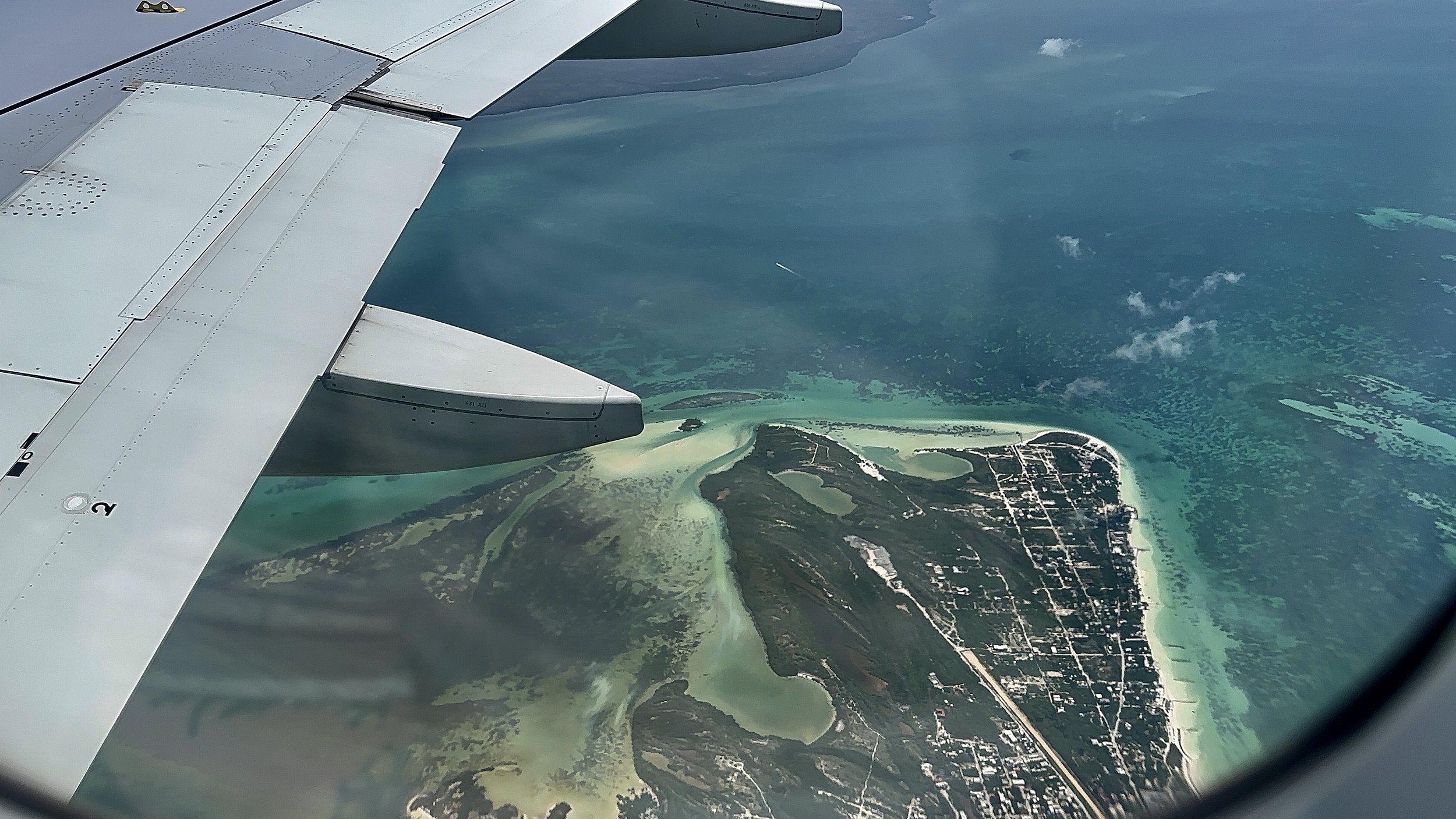 Aerial view from an airplane window showing a coastal landscape with turquoise water, sandy beaches, green land, and a visible plane wing.