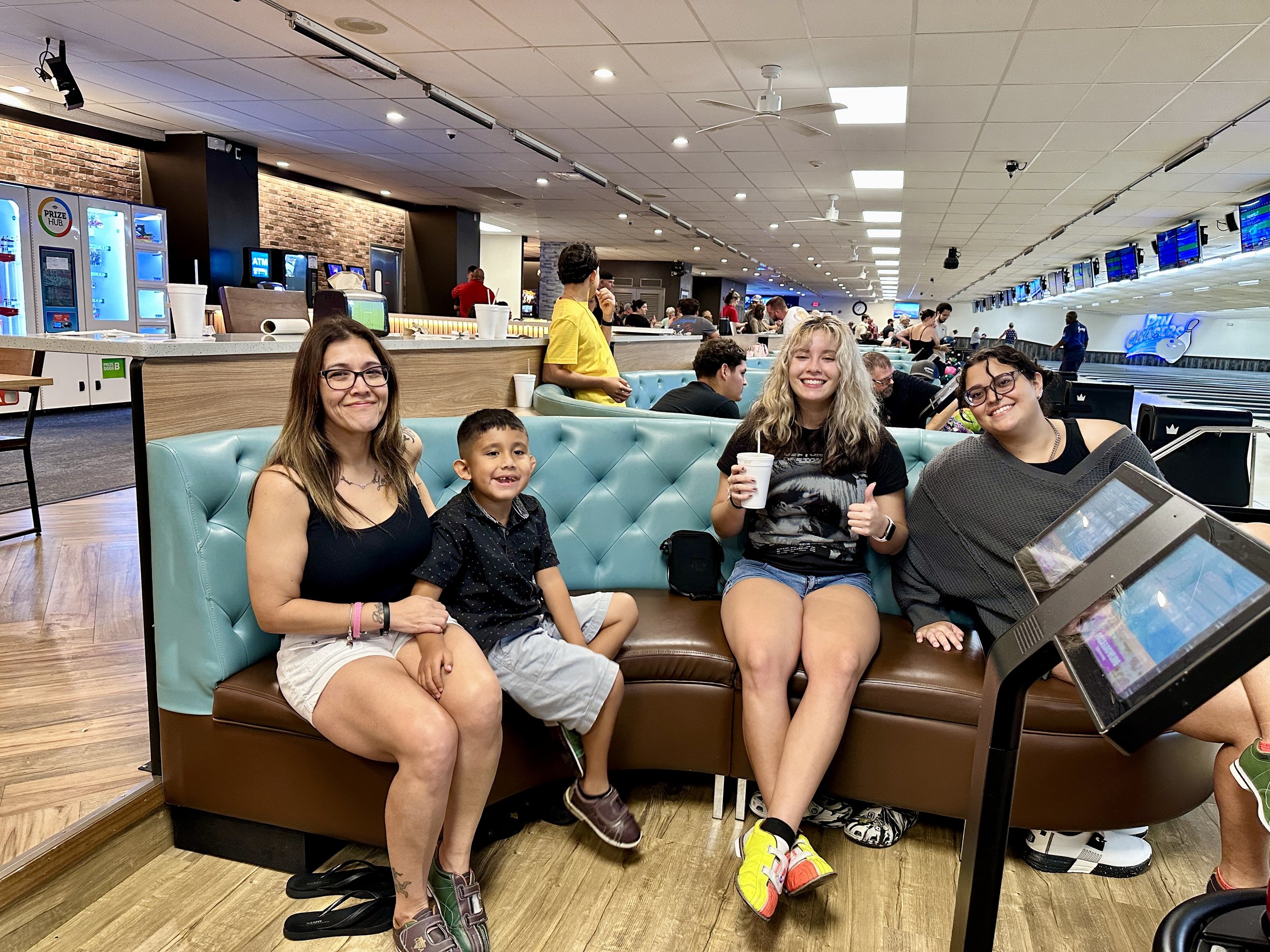 Group of people sitting on a leather couch at a bowling alley with lanes and screens in the background, enjoying a casual outing.