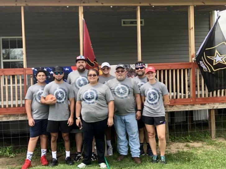 Group of people in matching t-shirts posing outdoors in front of a porch with flags, including a U.S. Army flag. One person holds a football.