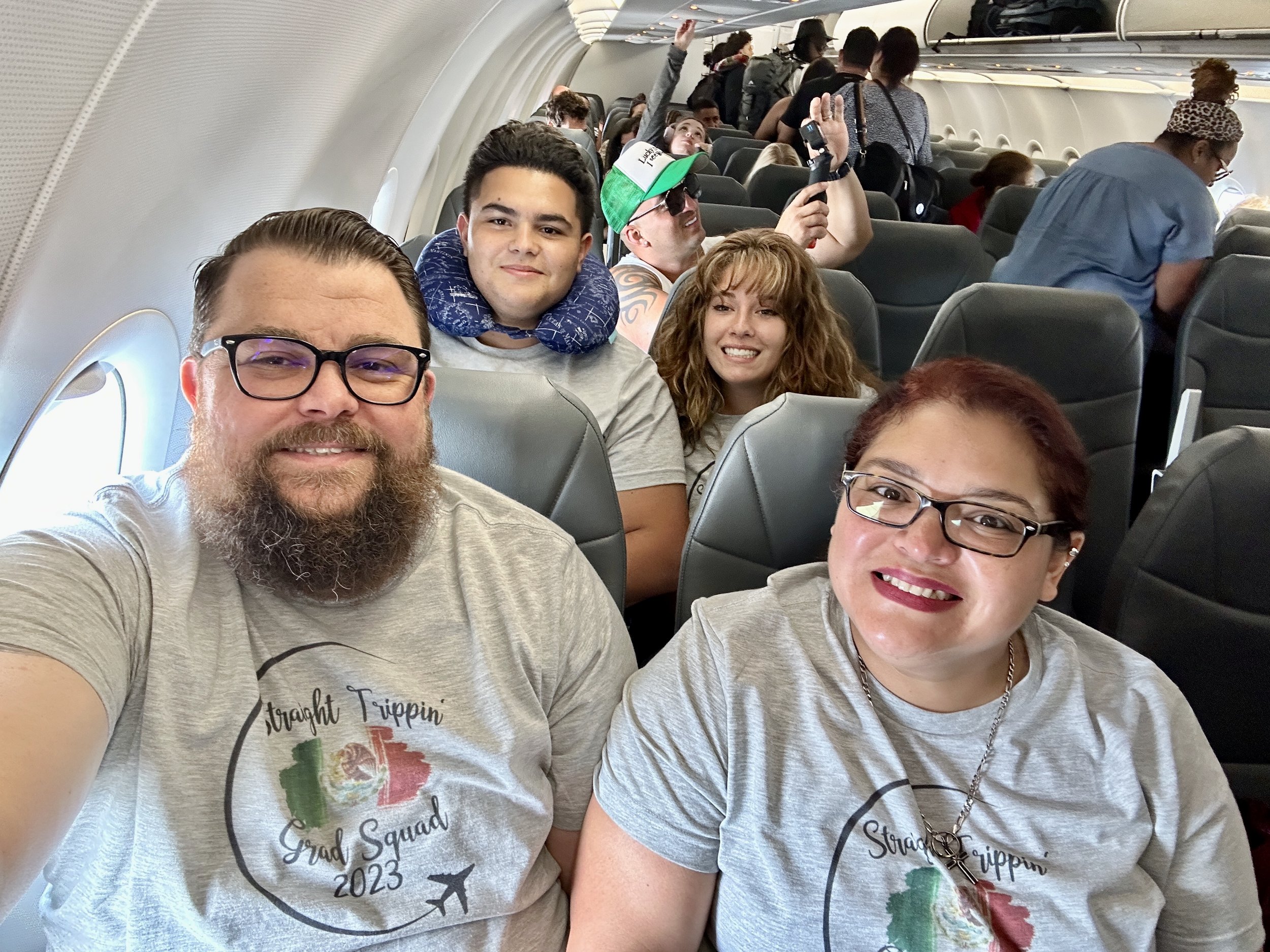 Group of people seated on an airplane, wearing matching "Straight Trippin’ Grad Squad 2023" t-shirts. Some are smiling at the camera while others are engaged in different activities.