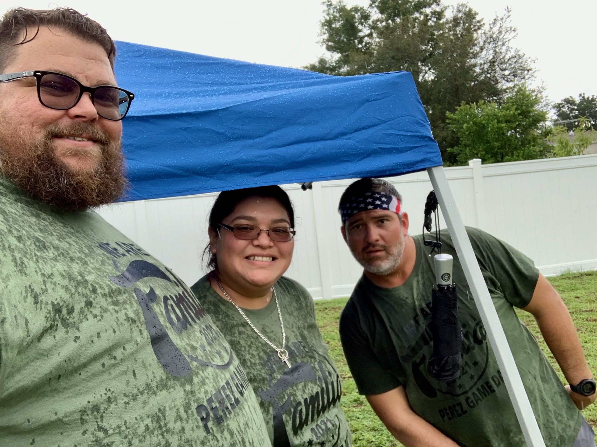 Three people in green shirts standing together under a blue canopy, outdoors on a rainy day, with a white fence and trees in the background.