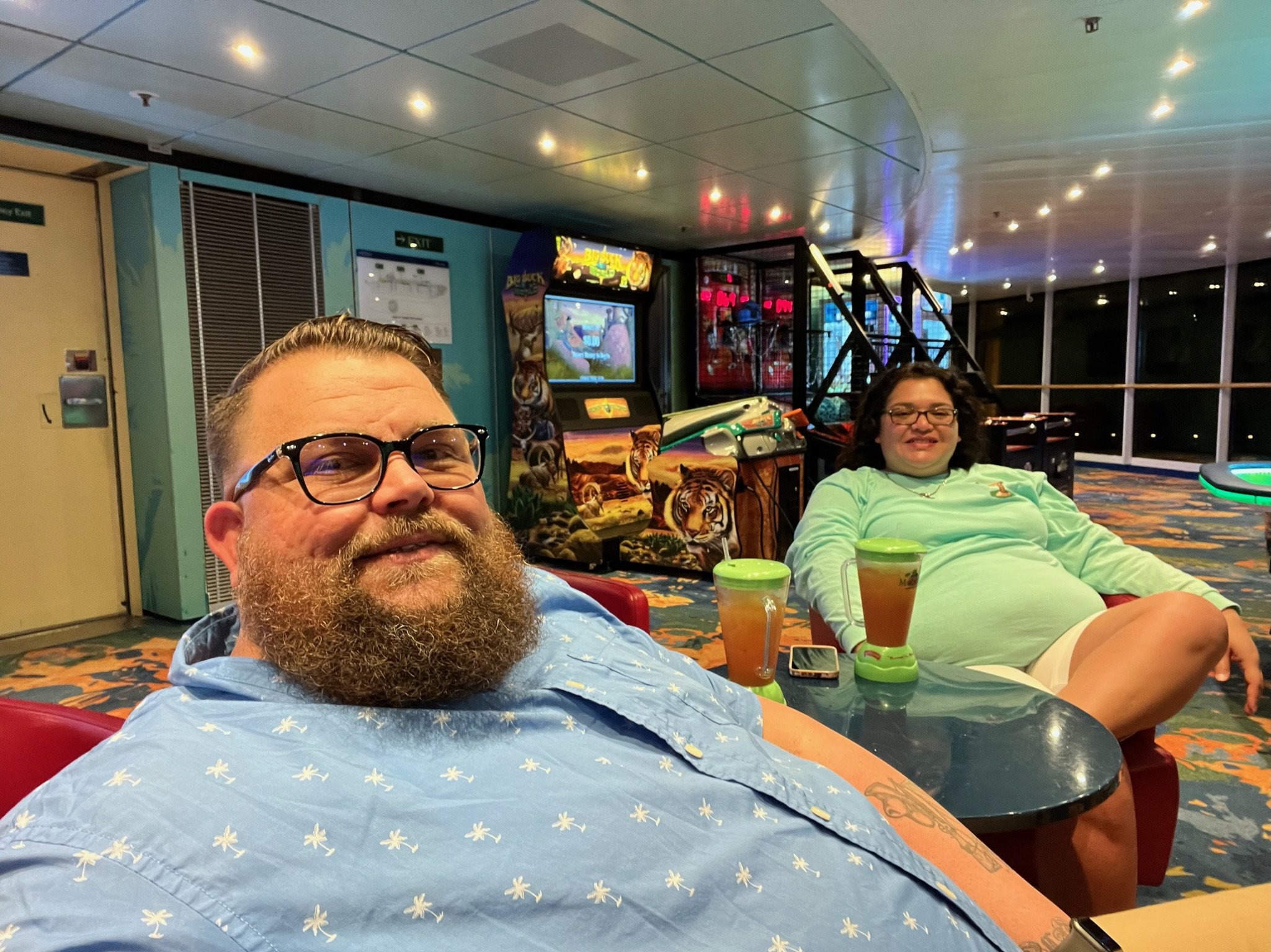 Two people sitting in a colorful recreational area, with arcade games and tables. They are relaxing with drinks in plastic cups. The area is well-lit and features vibrant decor.