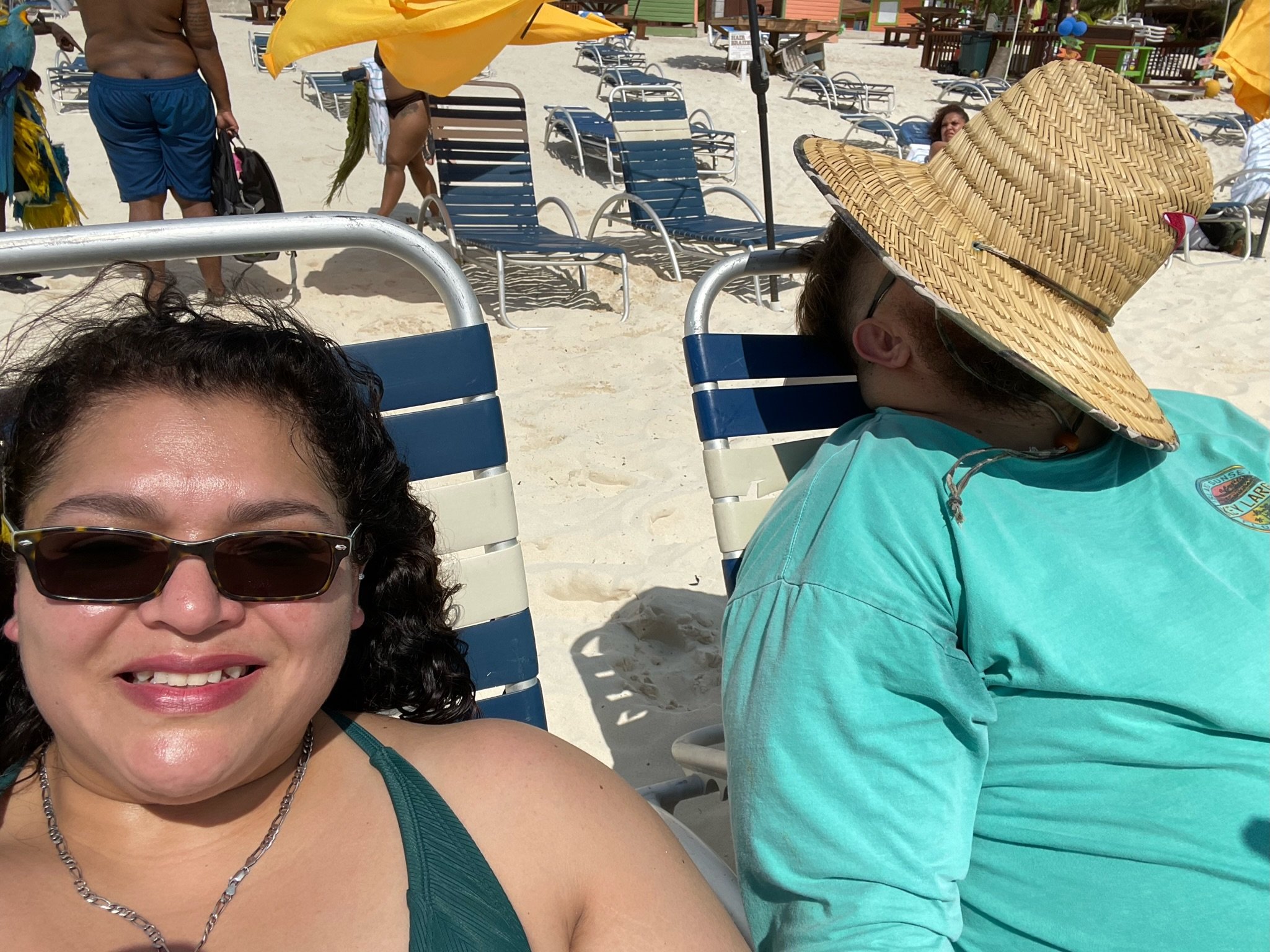 People relaxing on beach chairs at a sandy beach. A woman is smiling at the camera wearing sunglasses, while a man beside her is resting with a straw hat covering his face. Other people and beach chairs visible in the background.