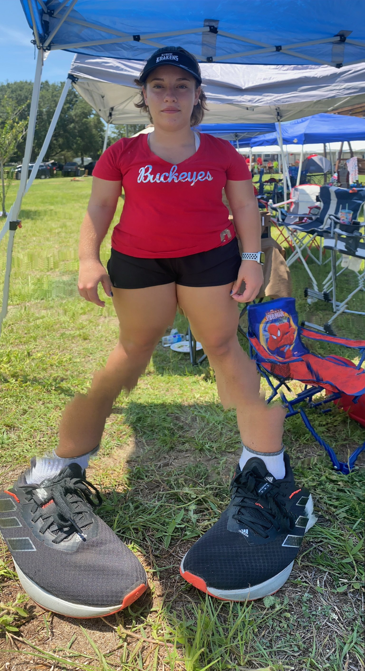 A person wearing a red "Buckeyes" shirt and black shorts with oversized sneakers, standing outdoors under a canopy. There are chairs and tents in the background on a grassy field.