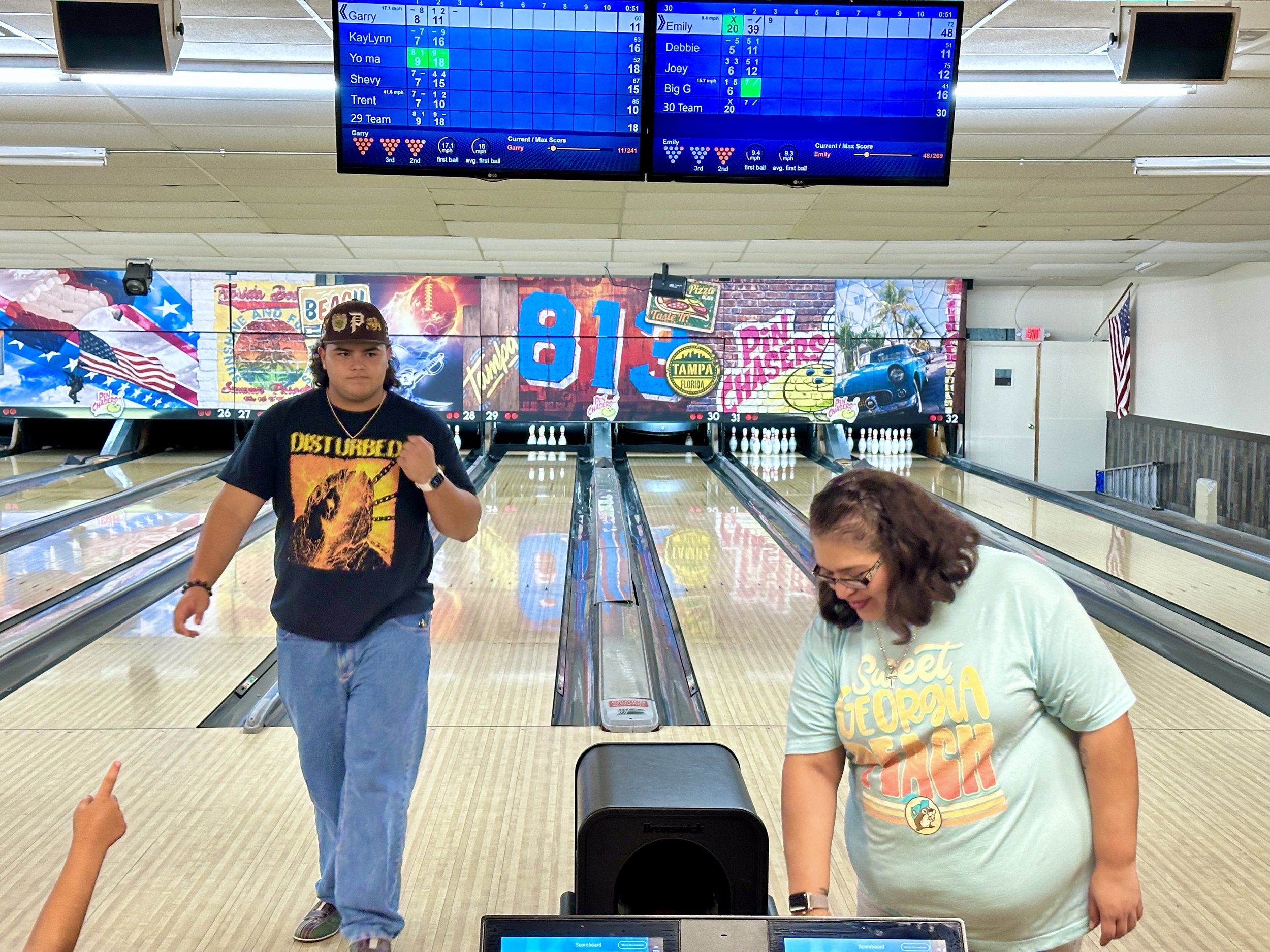 Two people in a bowling alley with lanes and an electronic scoreboard overhead, one person with a fist pump and the other looking down, colorful mural on the back wall.
