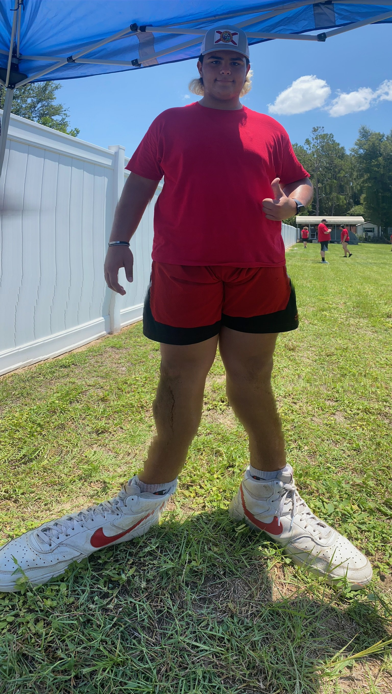 A person wearing a red shirt, shorts, and a cap standing on grass. The shoes appear oversized.