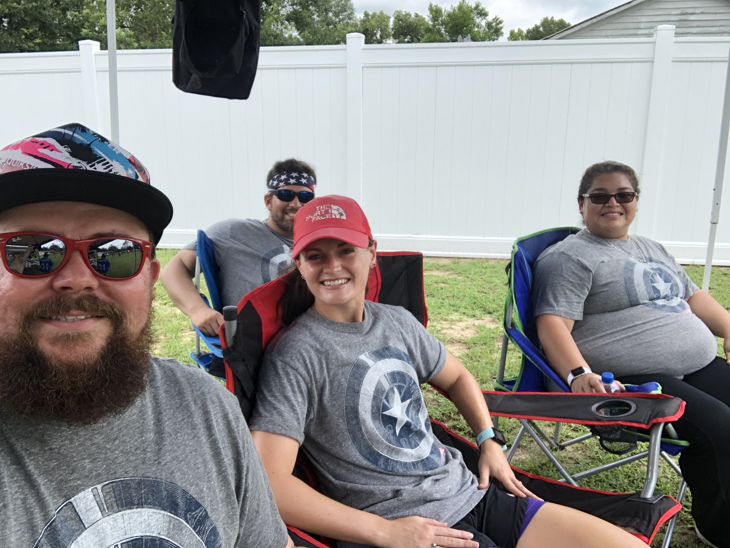 Group of four people sitting in outdoor chairs, all wearing matching gray shirts with a star emblem, in front of a white fence.