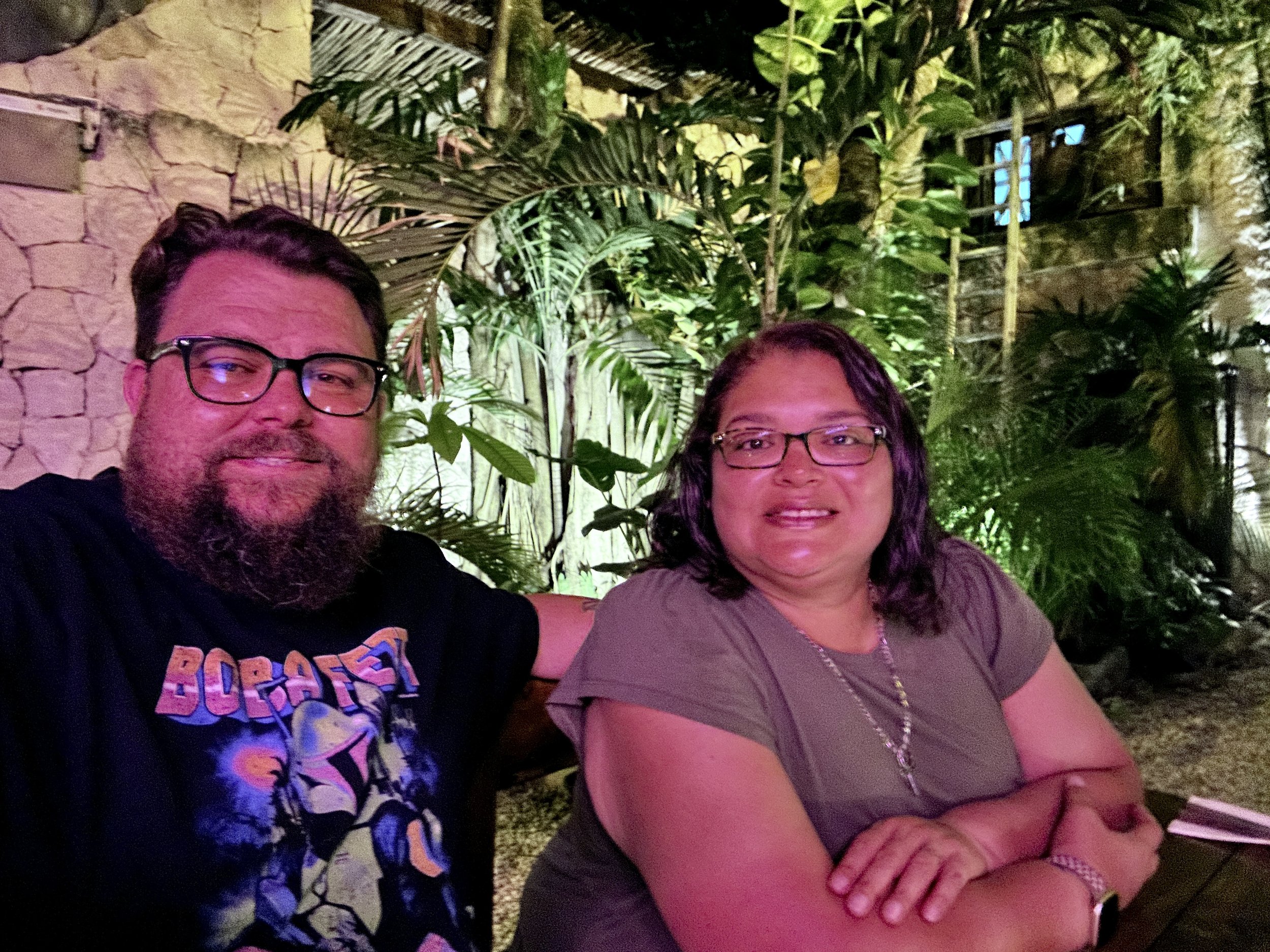 Two people sitting together, smiling, surrounded by plants and stone wall backdrop; one wears a Boba Fett T-shirt.
