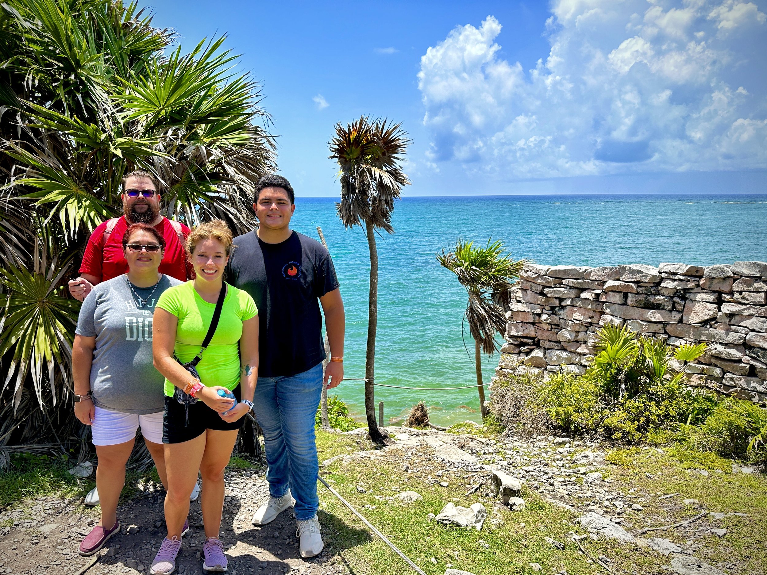Group of four people posing near tropical plants and stone ruins by the ocean.