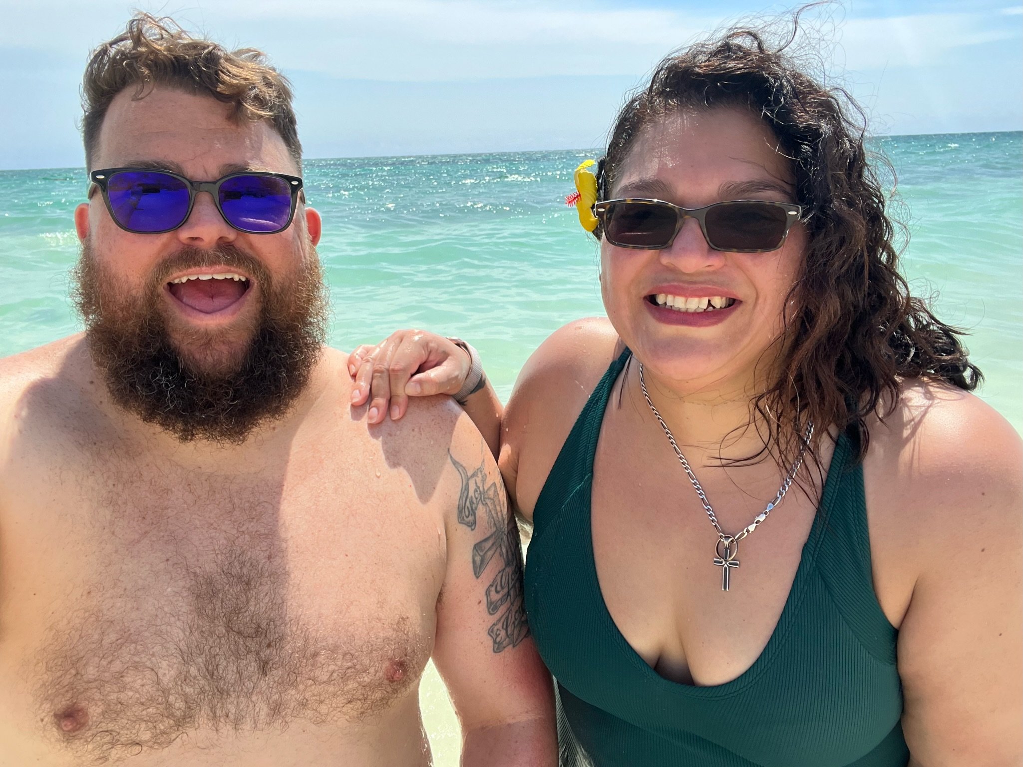 Two people smiling at the beach, wearing sunglasses and swimwear, with the ocean in the background.