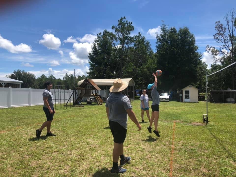 People playing volleyball outdoors in a grassy yard with a swing set in the background. One person is jumping to hit the ball.