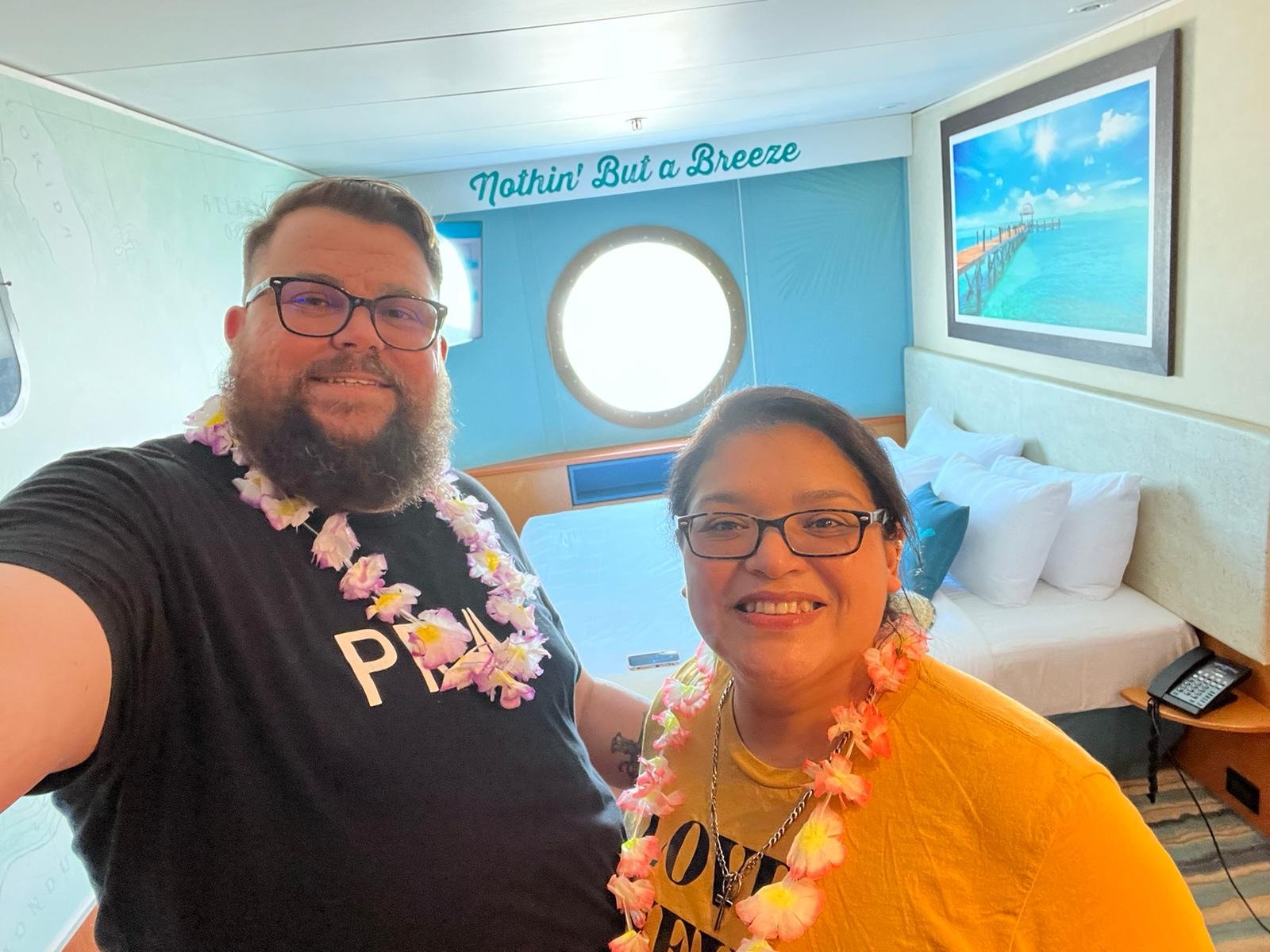 A smiling couple wearing floral leis inside a cruise ship cabin. The cabin has a round window, a bed with white pillows, and a wall decoration depicting a tropical scene. The phrase "Nothin' But a Breeze" is written above the window.